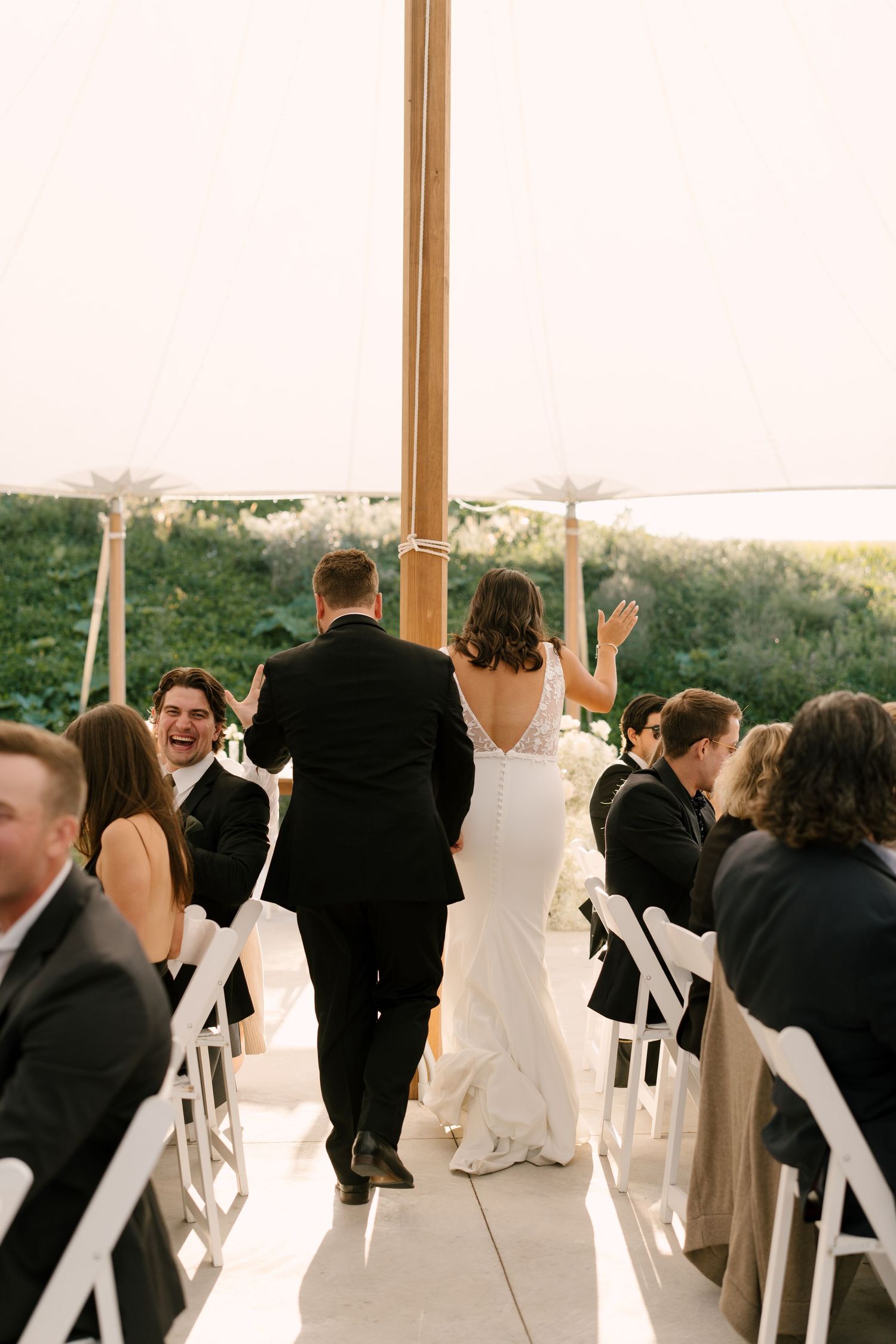 Bride and groom walk into wedding reception under elegant tent while guests cheer and celebrate.