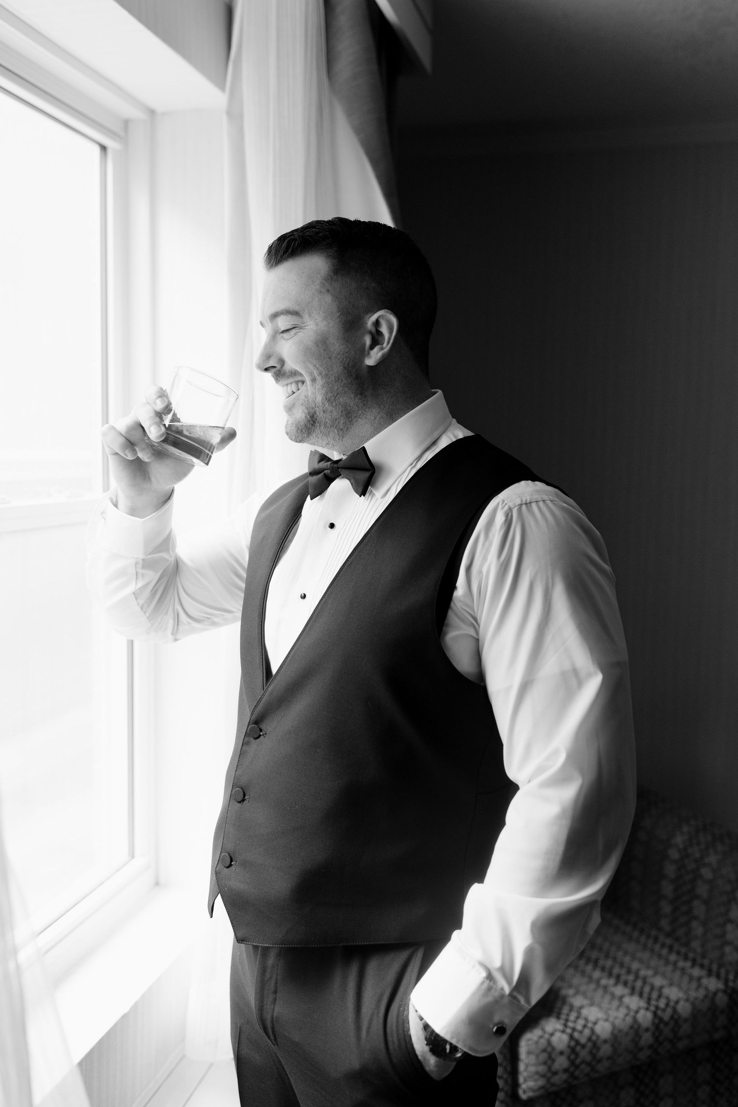 Groom in tuxedo vest and bow tie enjoying a glass of whiskey by a bright window in black and white.
