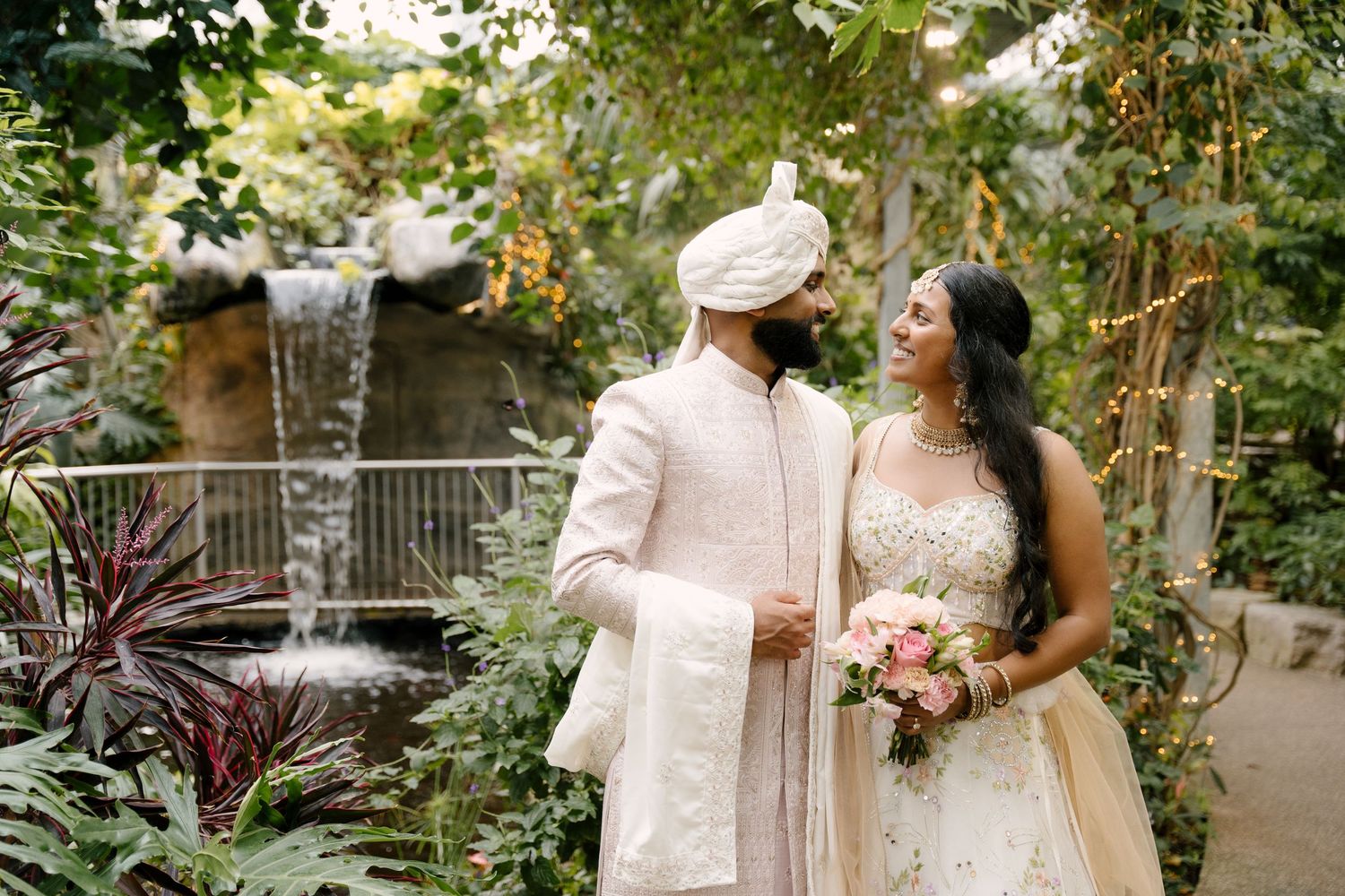 Indian couple in traditional wedding attire smile at each other in a lush greenhouse with a waterfall backdrop.