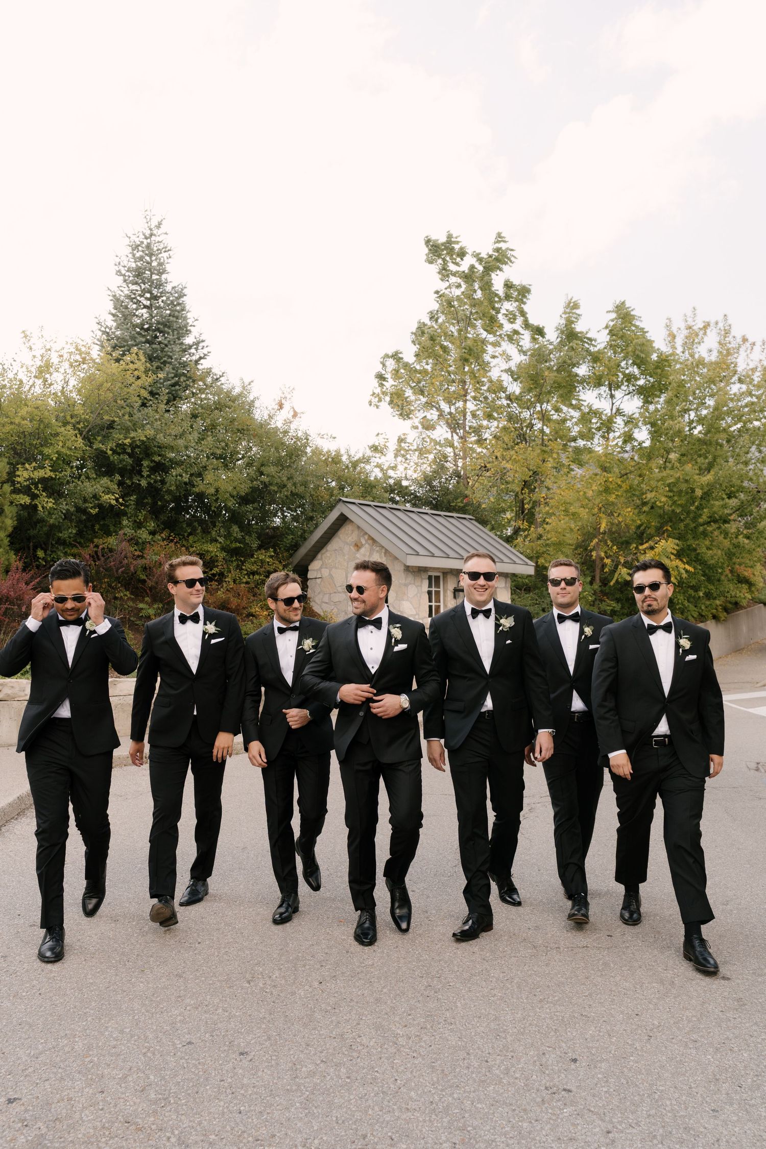 Seven groomsmen in black tuxedos and bow ties walk confidently outdoors at a wedding venue surrounded by trees at Cambridge Mill.