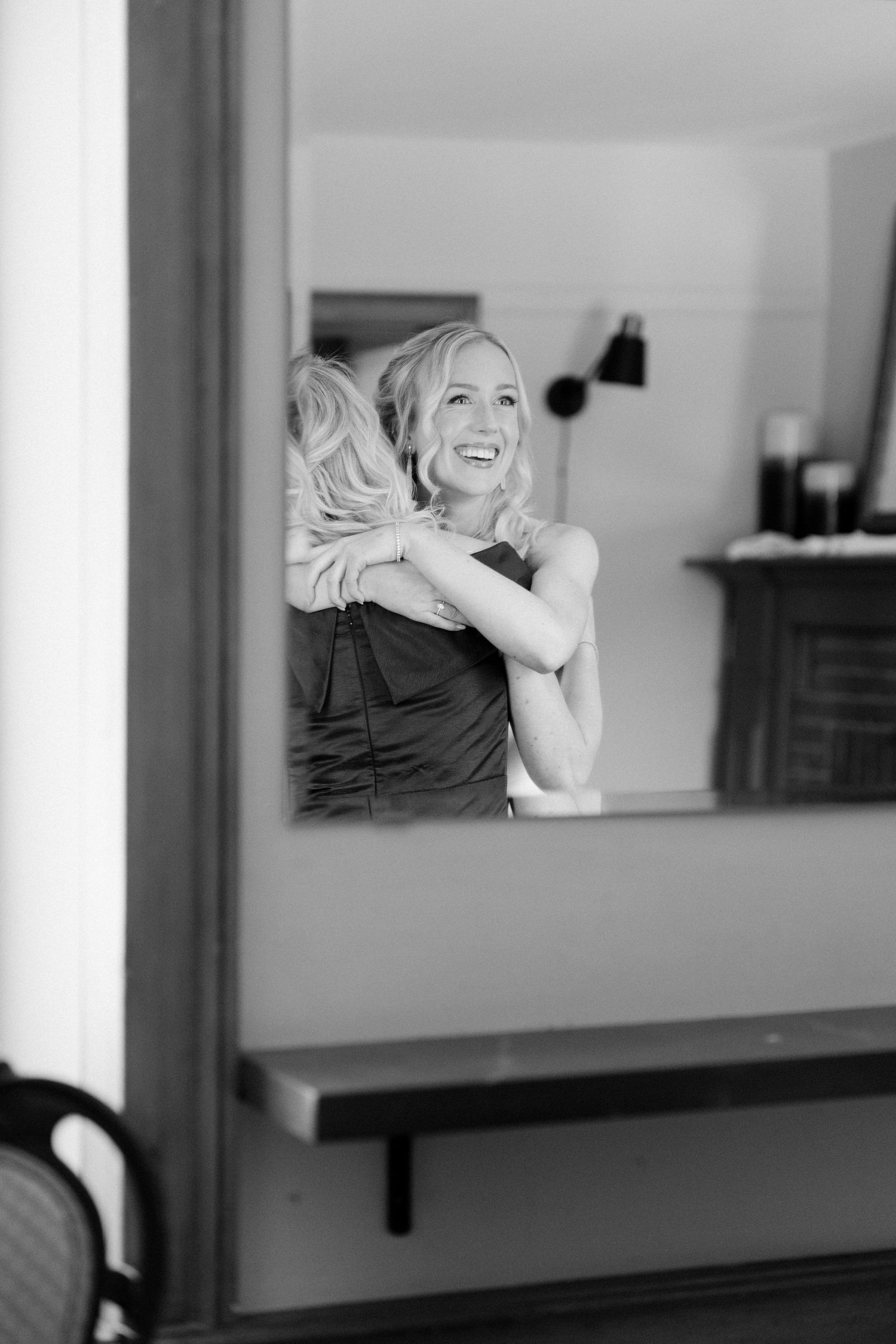 Joyful bride embracing bridesmaid captured in mirror reflection in black and white wedding preparation photo.