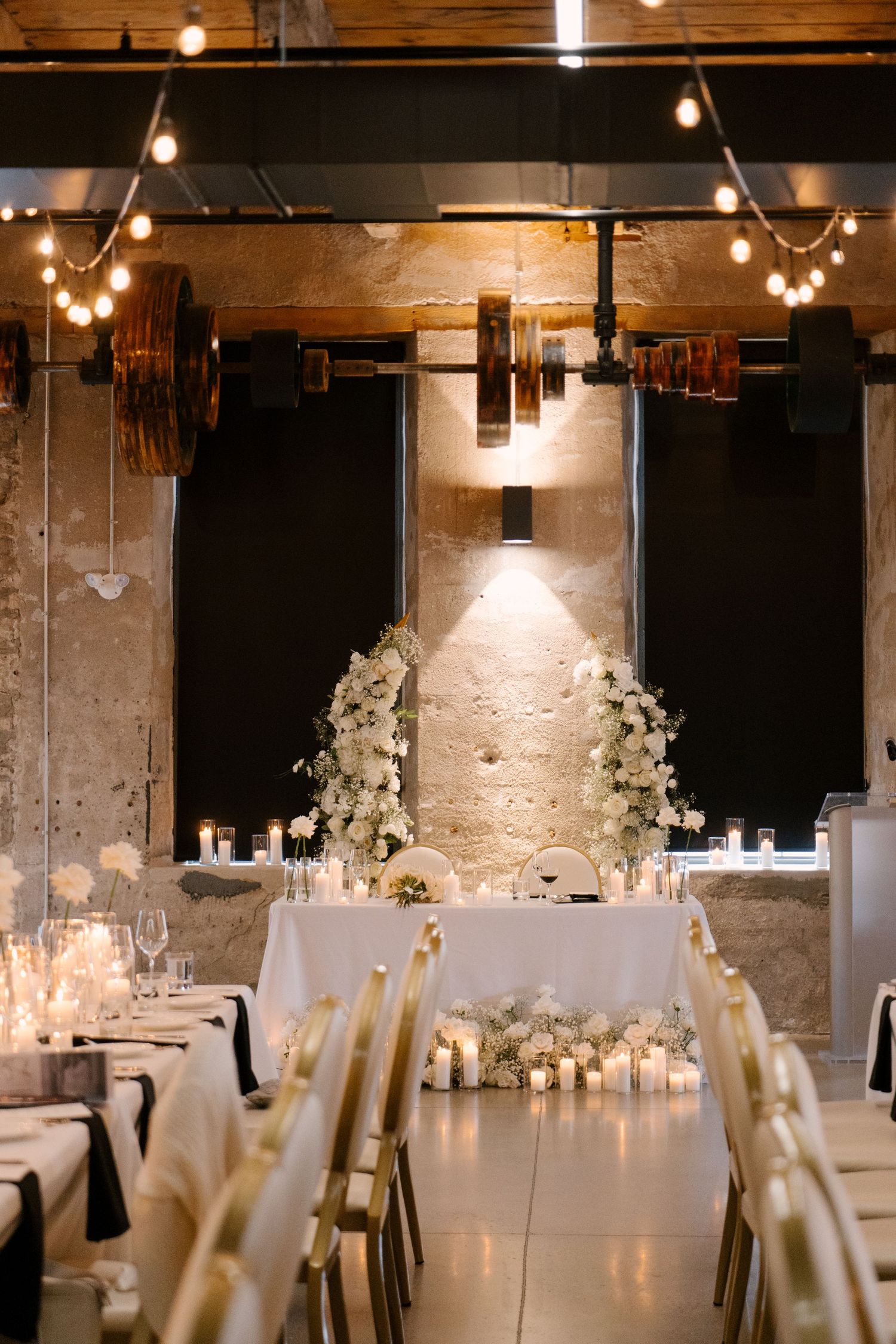 Tapestry Hall wedding venue with string lights, white floral sweetheart table arch, and glowing candles on long reception tables.
