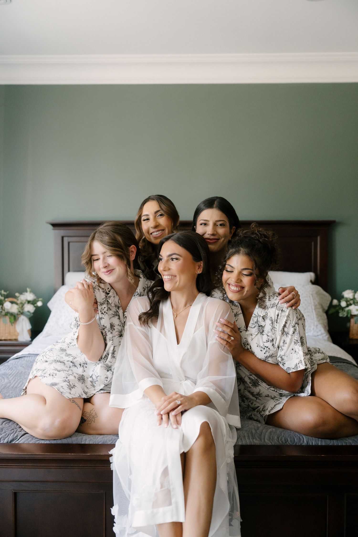 Bride in white robe poses with four bridesmaids in floral robes on a bed during wedding morning preparations.