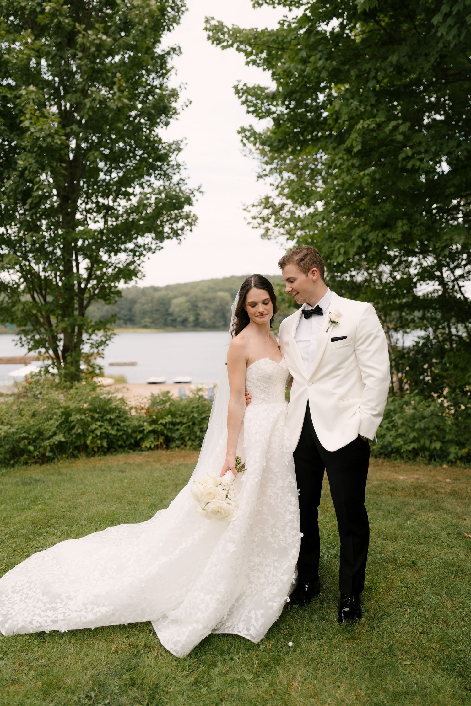 A bride in a lace gown and groom in a white tuxedo stand on a lawn by a lake surrounded by trees.