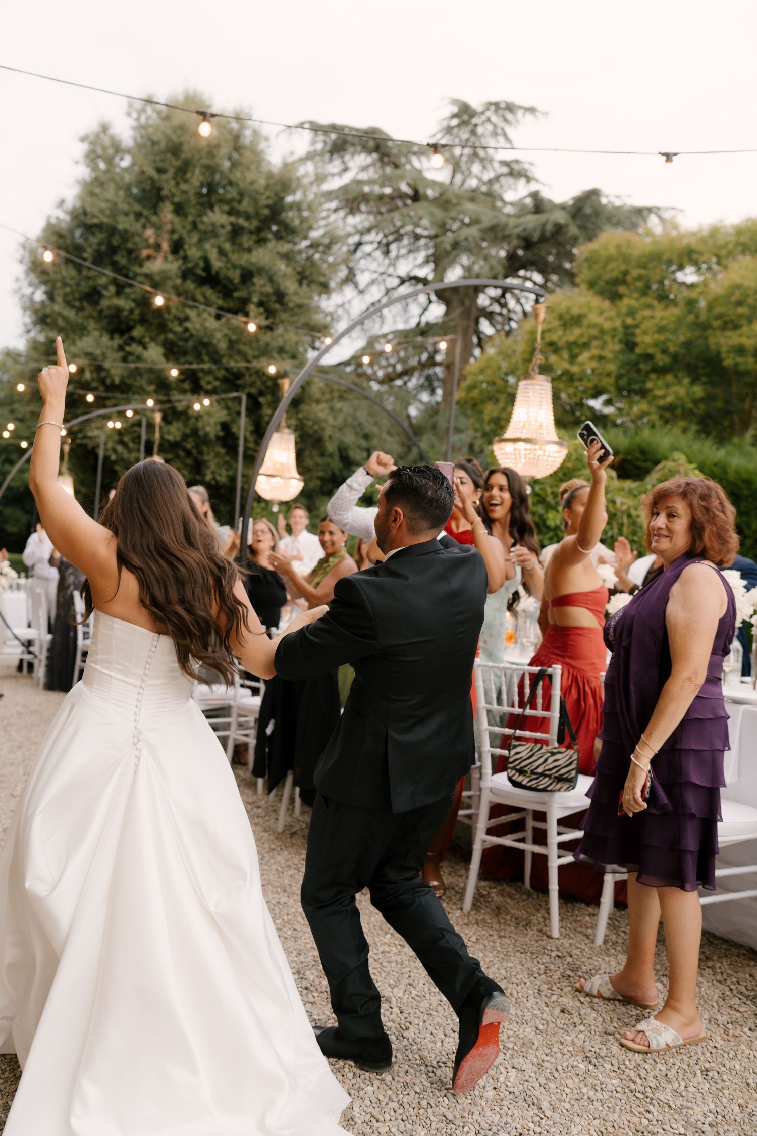 Bride and groom dancing joyfully with guests at outdoor Tuscan wedding reception at dusk.