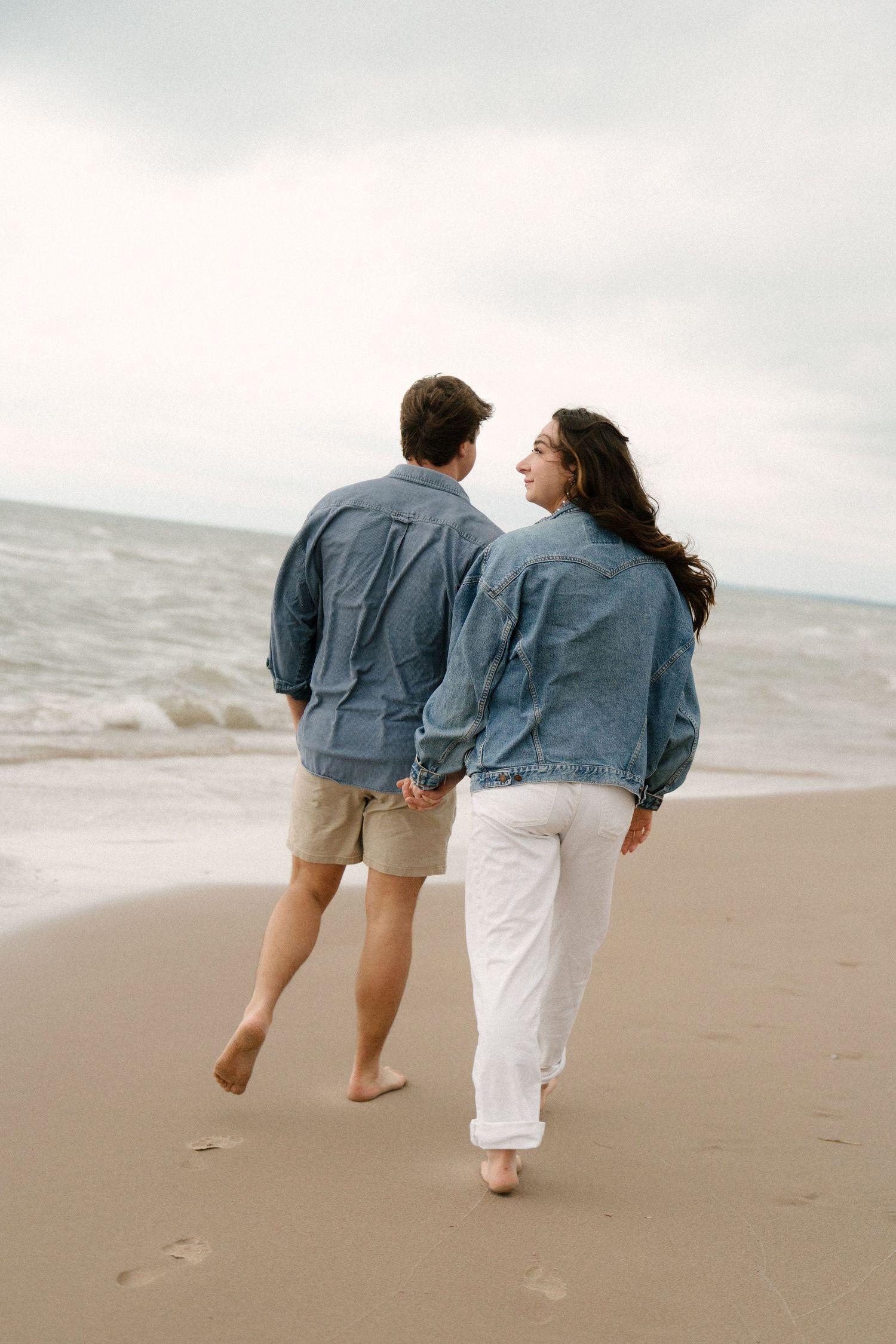Couple in matching denim jackets walking hand in hand along the shoreline leaving footprints in sand.