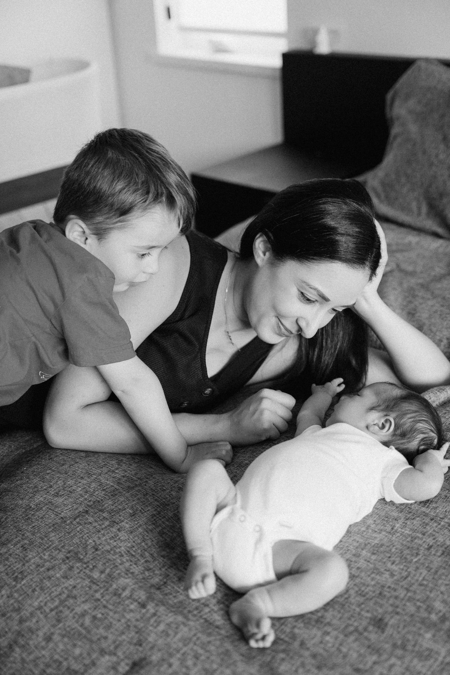 Black and white photo of mother and young son smiling at newborn baby on gray couch.