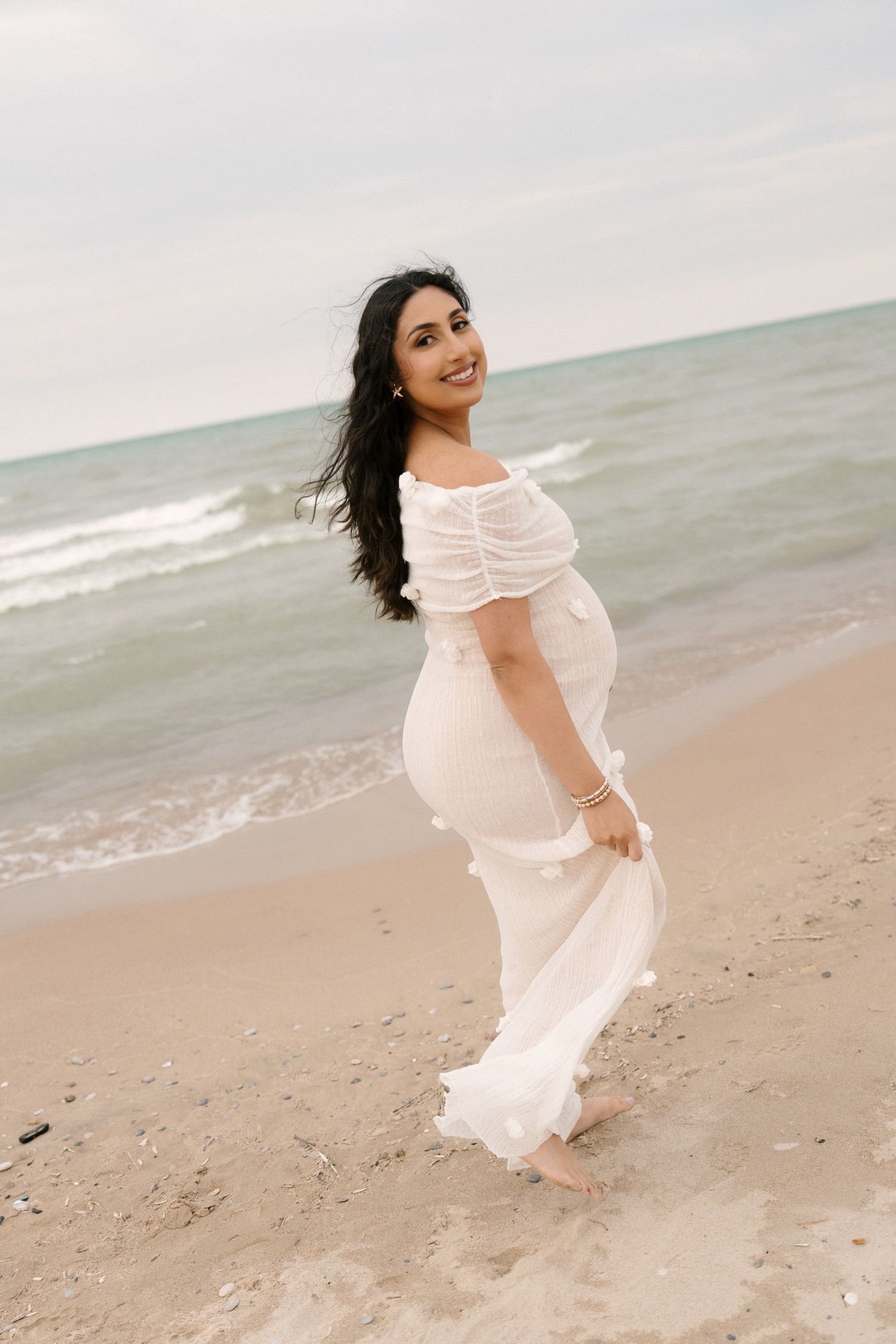 Pregnant woman in white off-shoulder gown walking on sandy beach with ocean waves behind her.