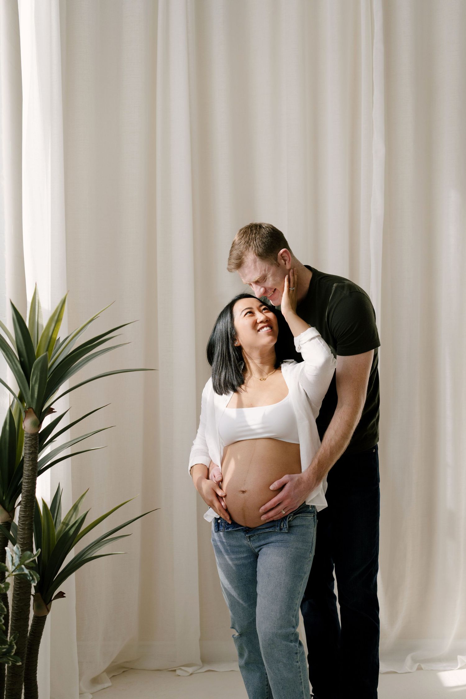 Man in dark shirt embraces pregnant woman from behind, both smiling, standing before white curtains.