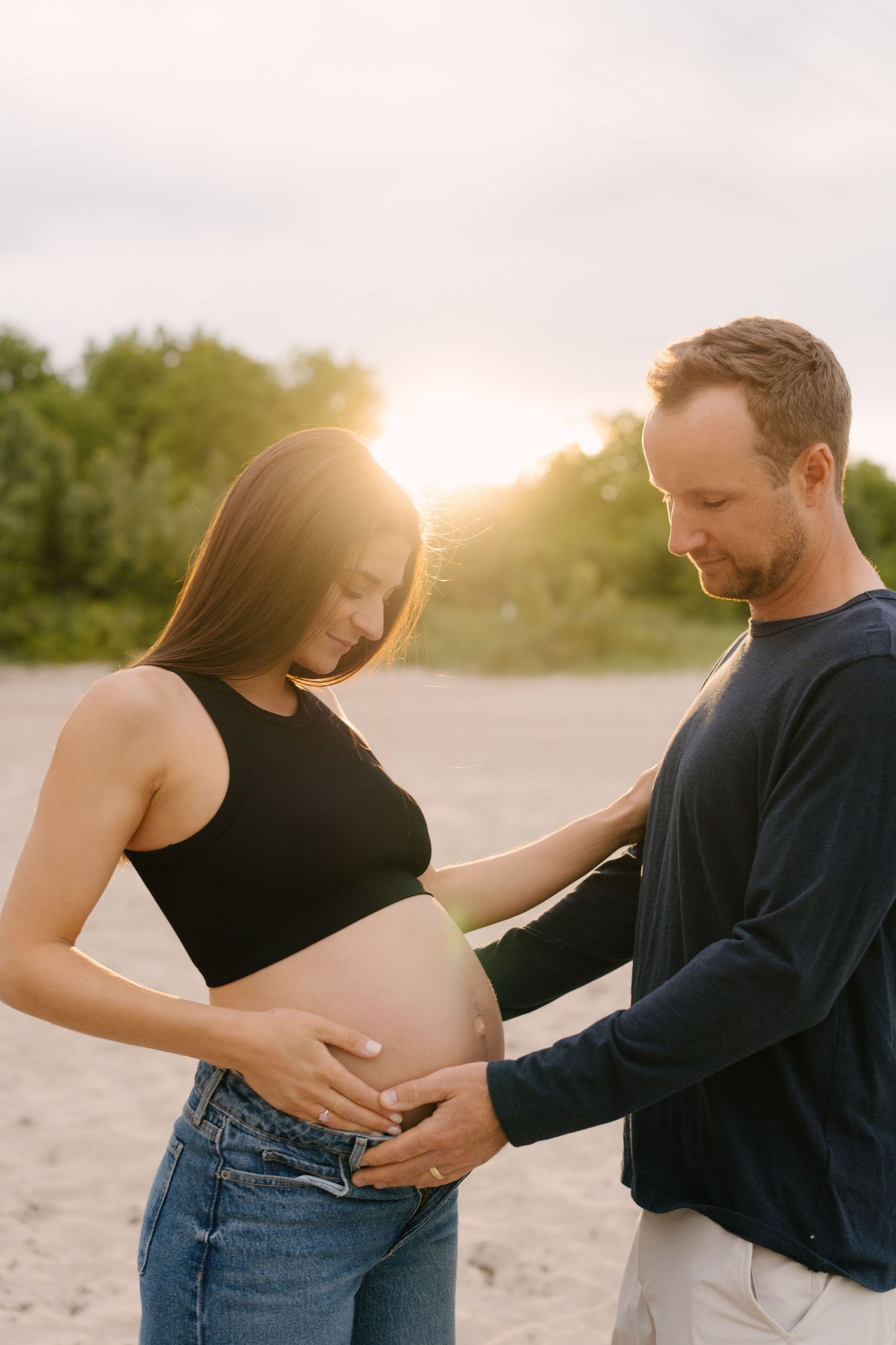 Man gently touches pregnant womans baby bump at golden hour on a sandy beach with glowing sunset behind them.