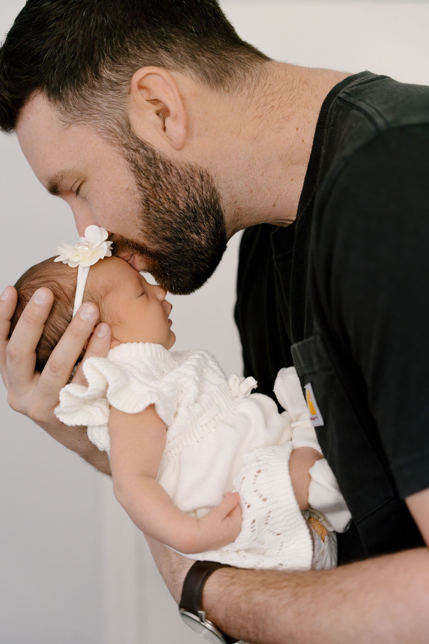 Loving bearded father gently kissing his sleeping newborn baby girl dressed in white lace outfit.