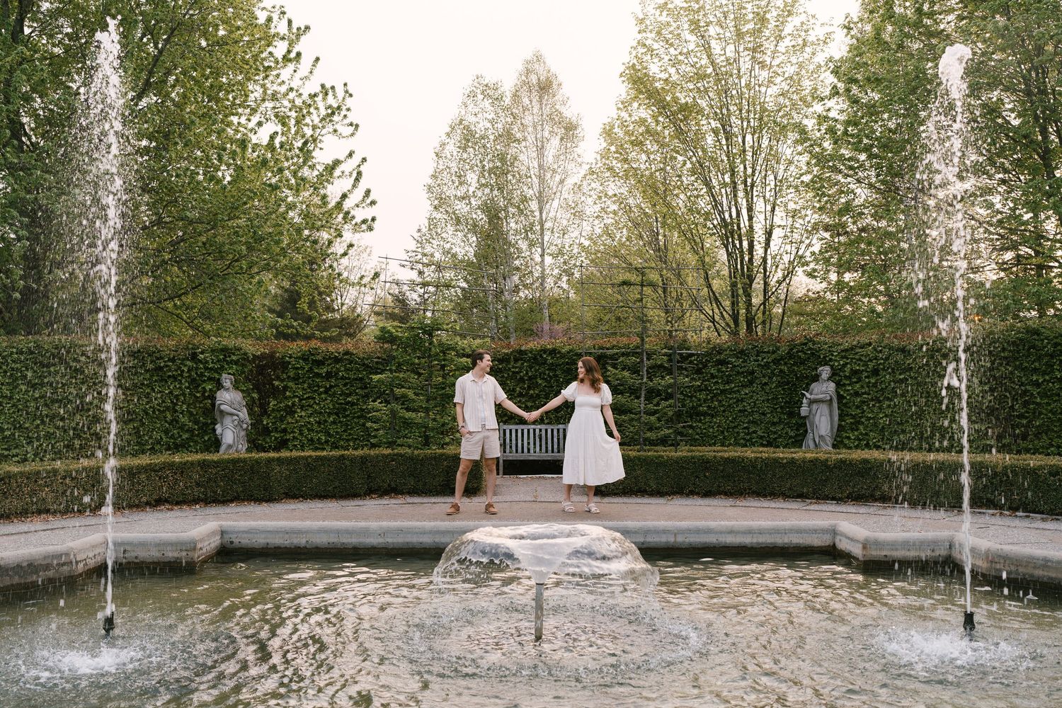 Couple holding hands beside a decorative fountain in an elegant formal garden with tall green hedges.