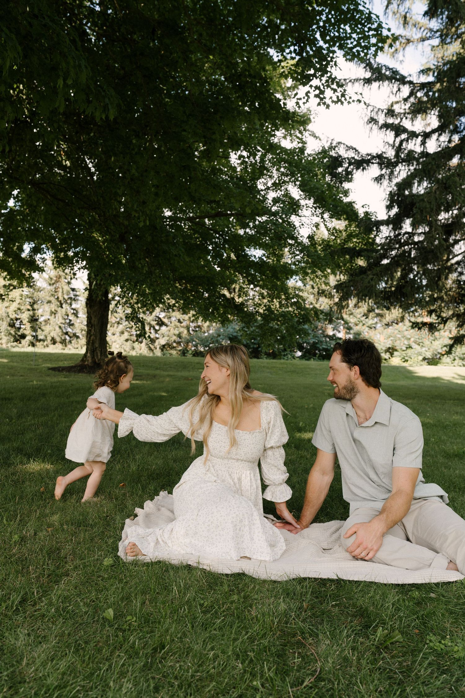 A family relaxes on a blanket under a large tree as their toddler daughter runs toward her mother.