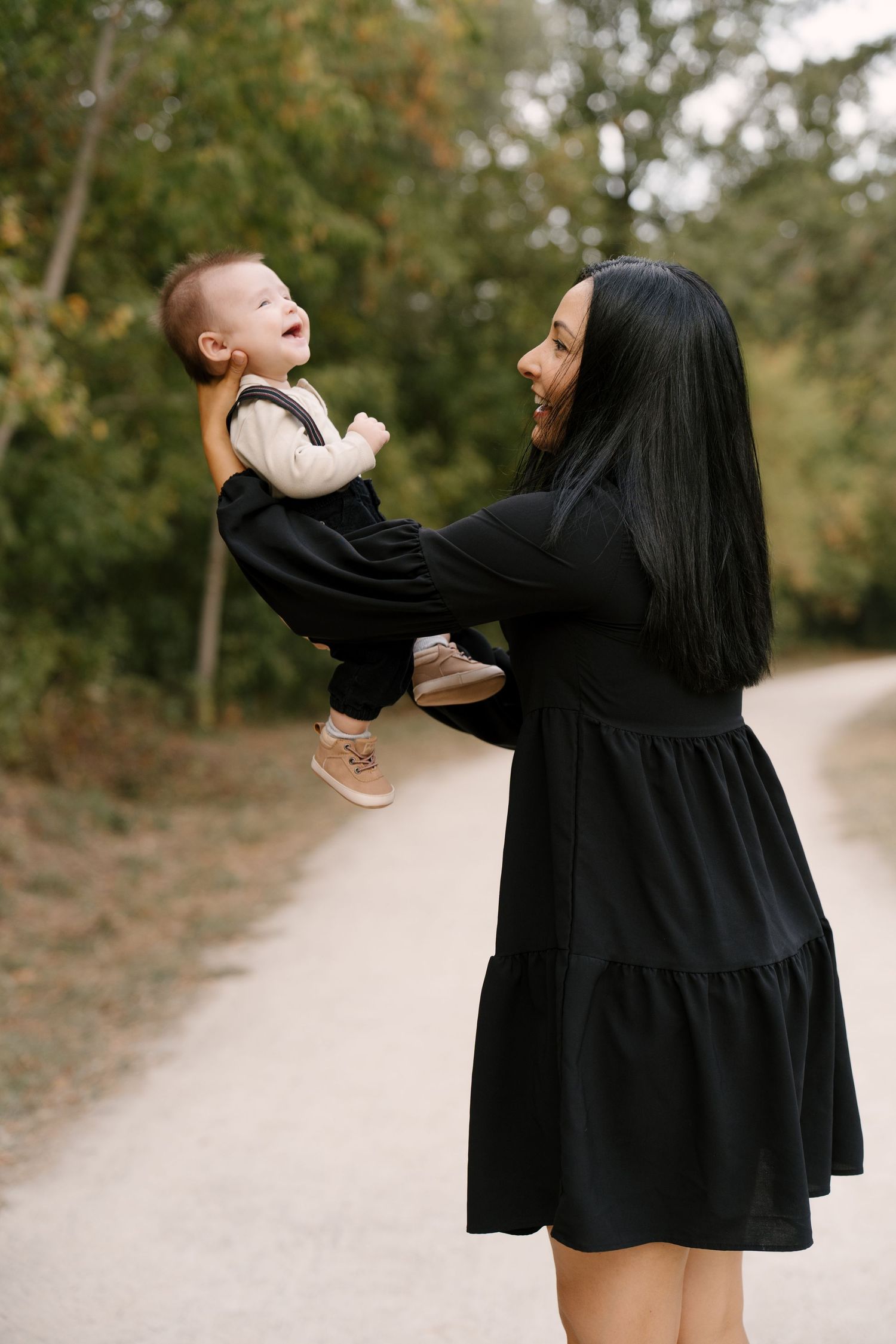 Smiling mother in black dress lifts her laughing baby in the air on a scenic outdoor path.