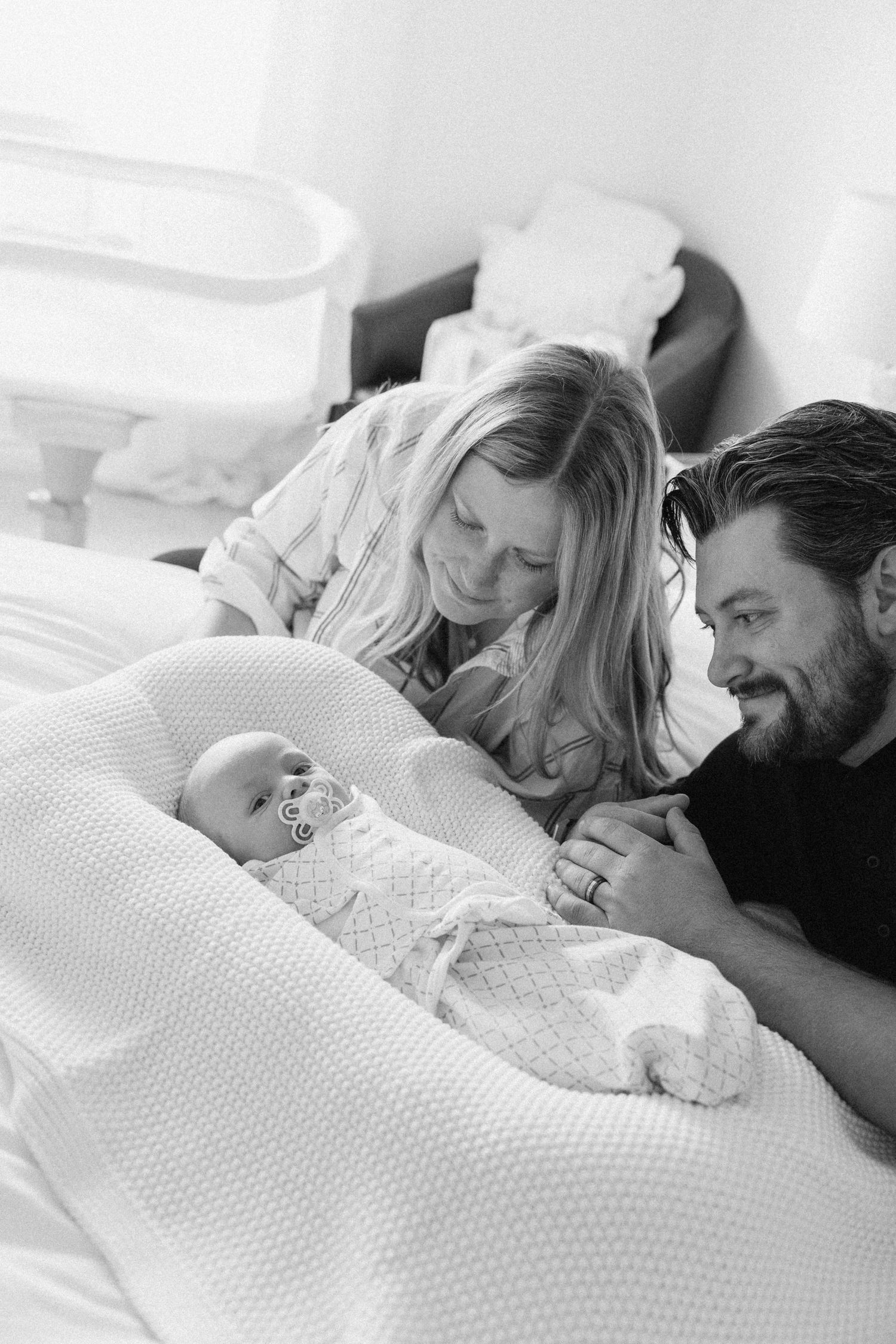 Black and white photo of new parents lying on bed smiling at their newborn baby in a snuggle nest.