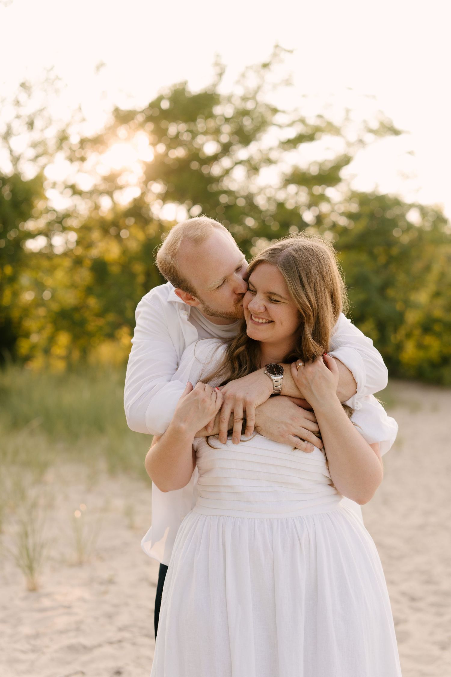 Man in white shirt embracing woman in white dress from behind on sandy beach at sunset.