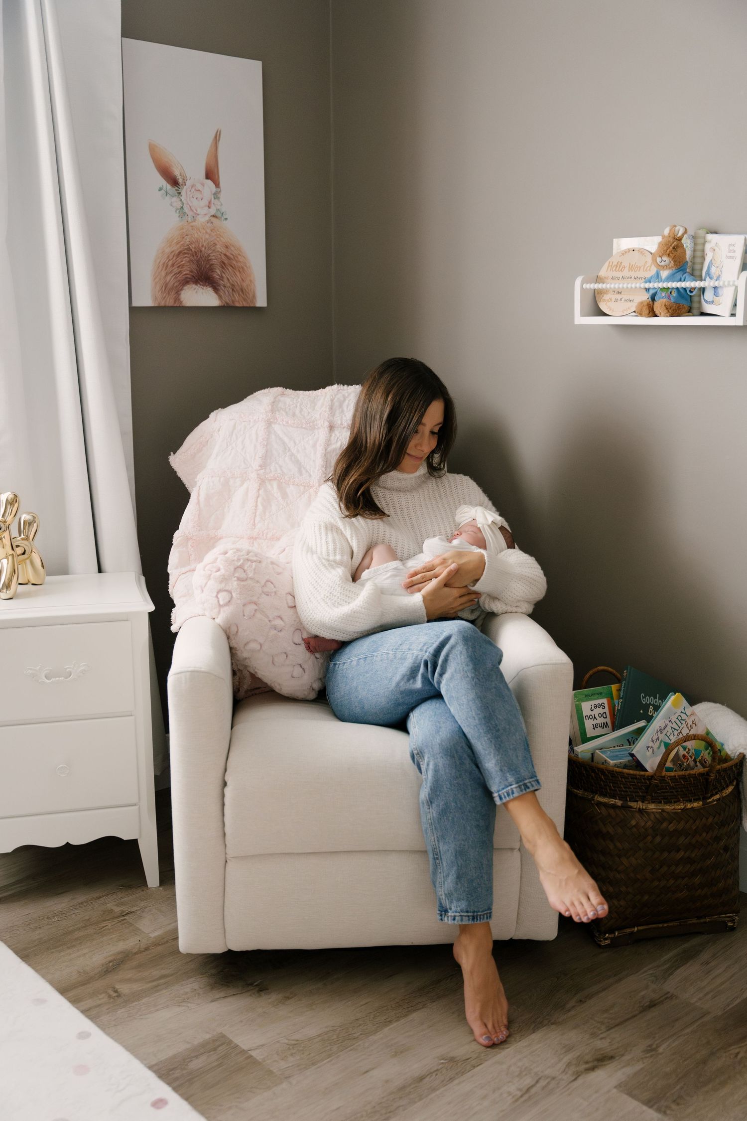 New mother in white sweater and jeans sitting in nursery glider chair holding sleeping newborn baby.