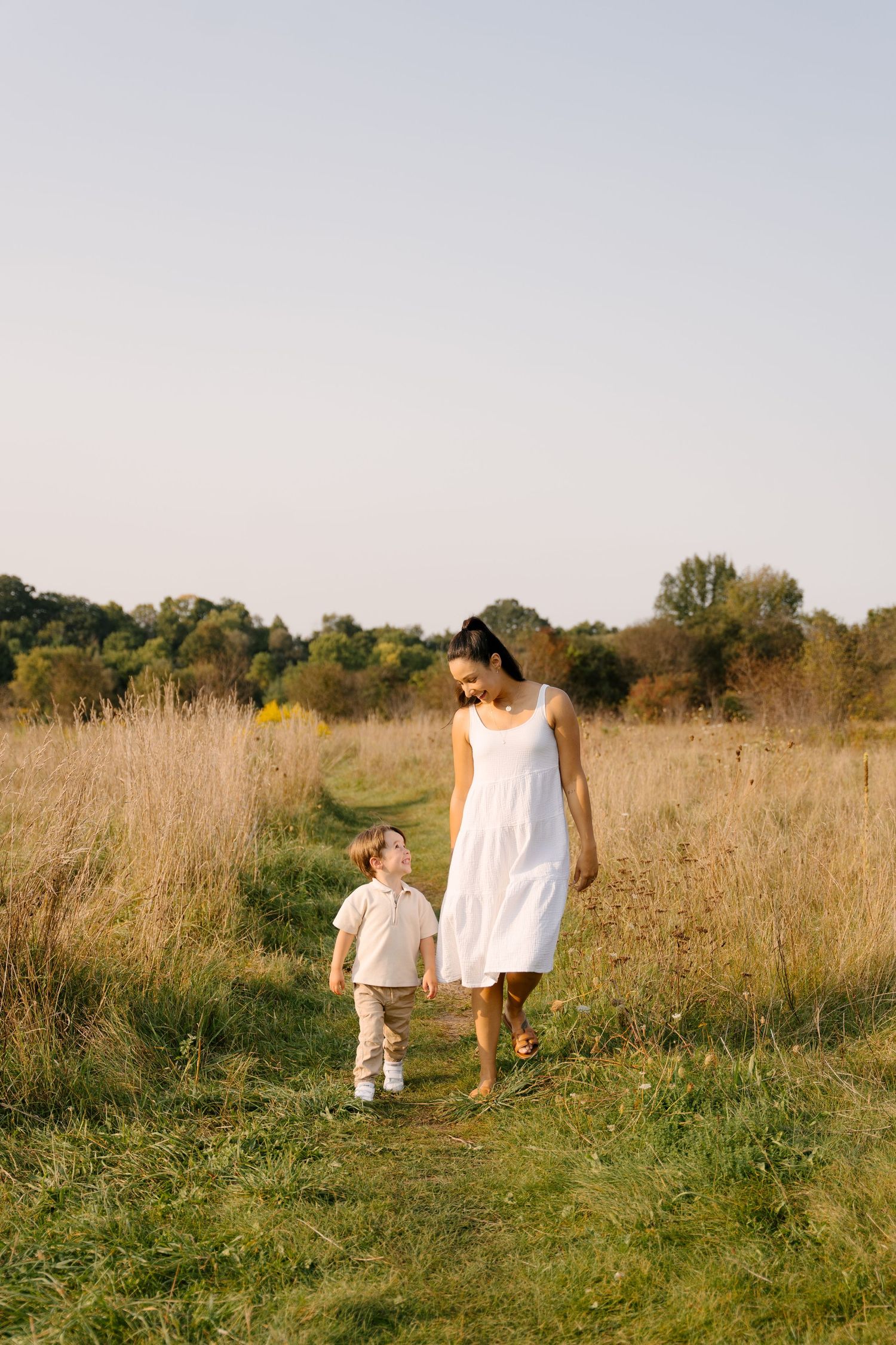 Mother in white dress walks with toddler son along a path through a golden meadow on a sunny day.