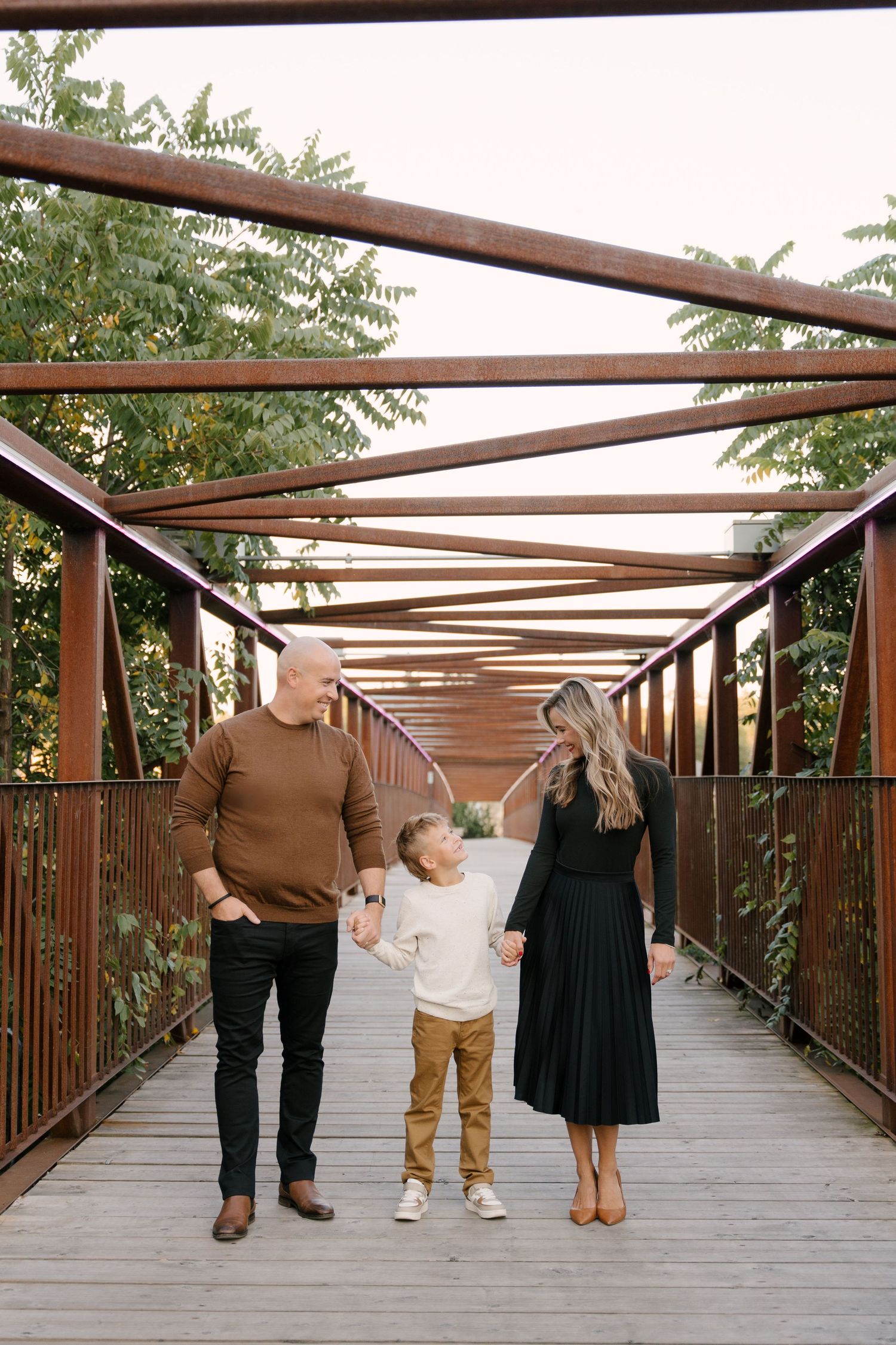 Happy family of three holding hands while walking across a rustic iron bridge.
