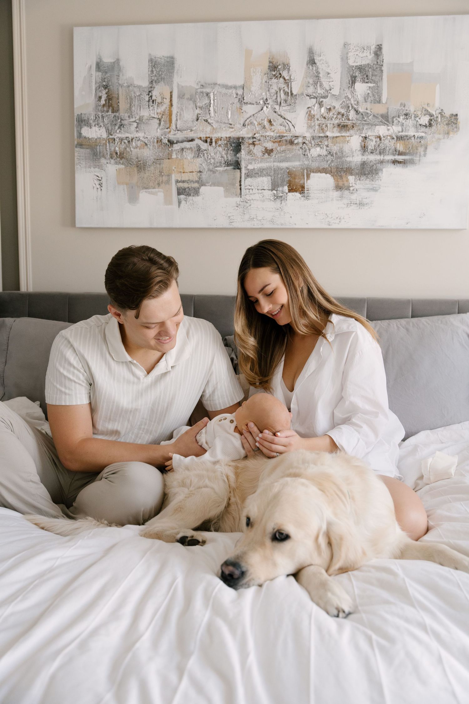 A young couple sits on a white bed with their newborn baby and golden retriever dog in a bright bedroom.