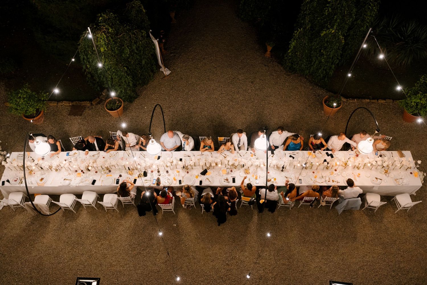 Aerial view of long wedding reception dinner table with guests seated outdoors at night.