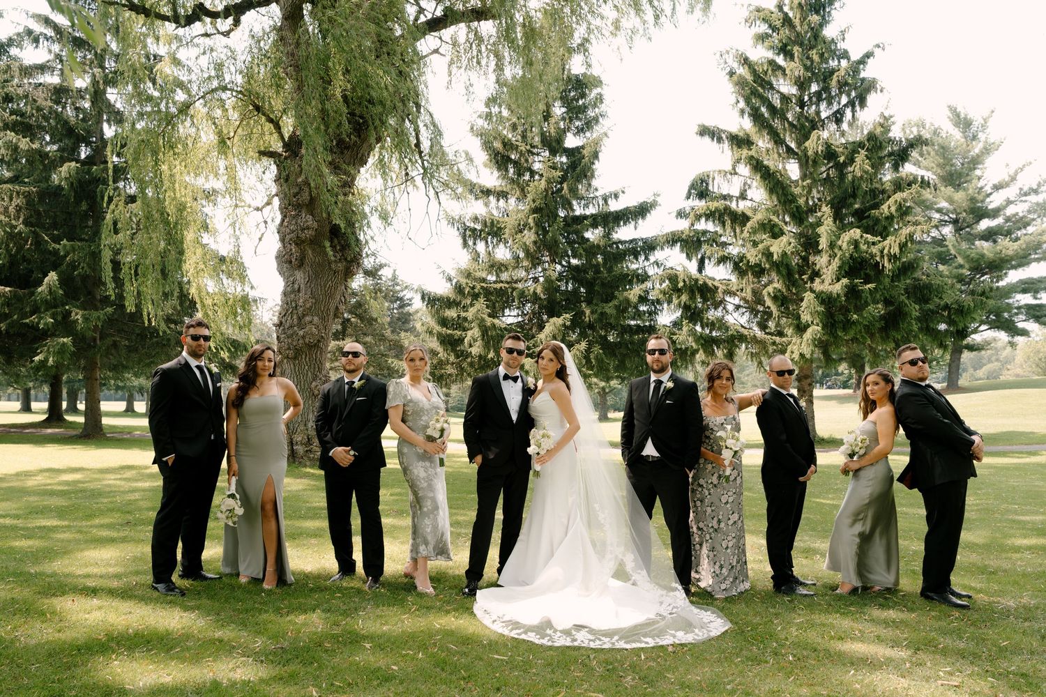 Wedding party poses outdoors on lush green lawn with tall pine trees, bride in white gown surrounded by groomsmen and bridesmaids.
