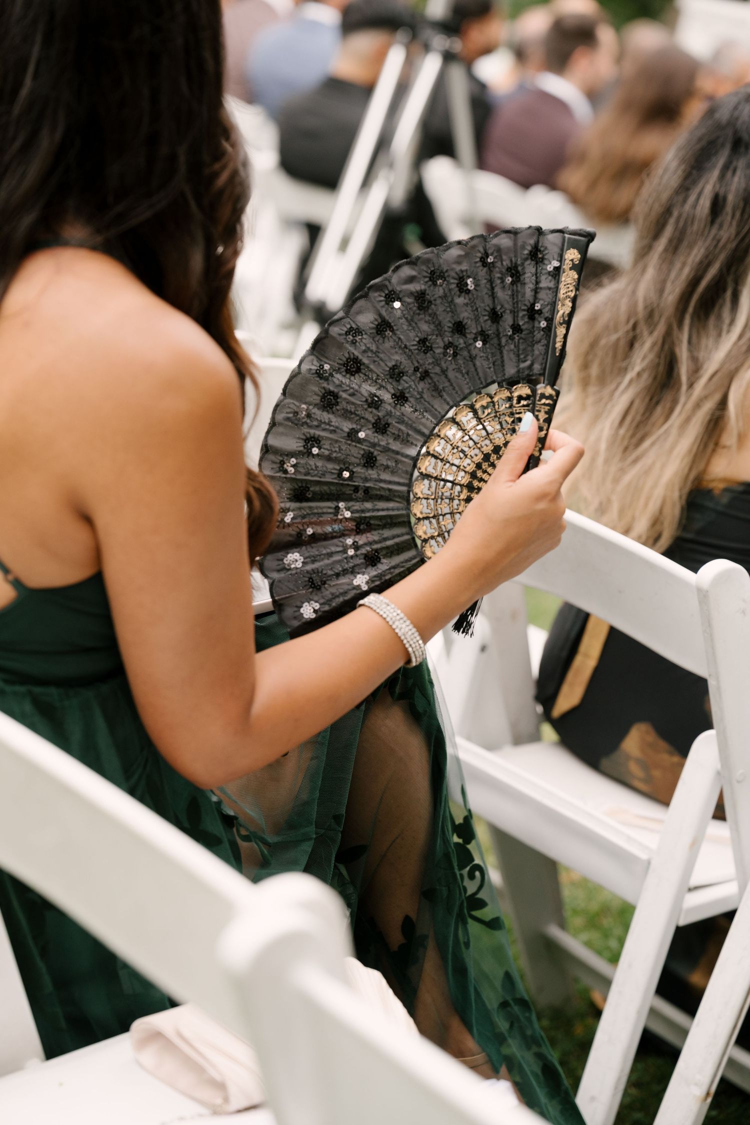 Wedding guest in green dress holding an ornate black and gold decorative hand fan during an outdoor ceremony.