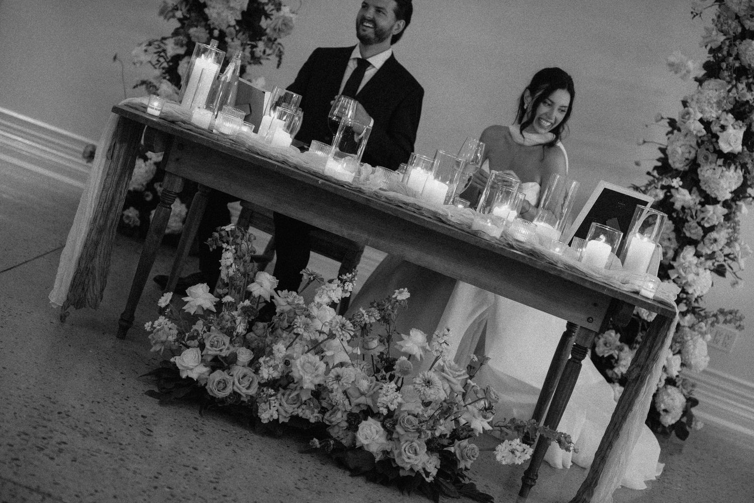 Black and white photo of bride and groom seated at floral-decorated sweetheart table with candles at Roseville Estates wedding reception.