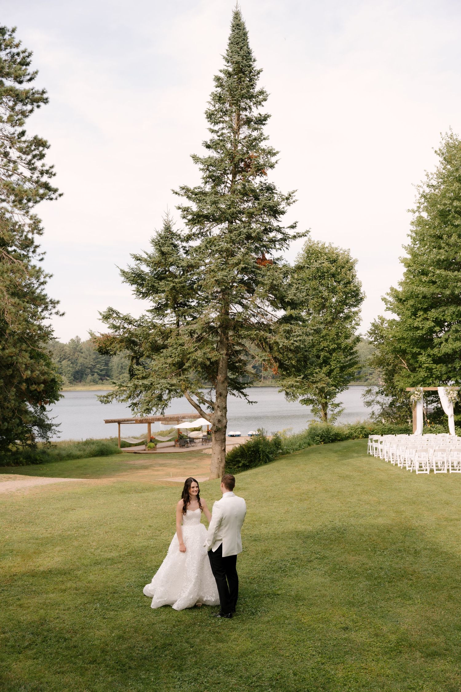 Bride and groom hold hands on green lawn near tall pine tree with lake and white chairs in background.
