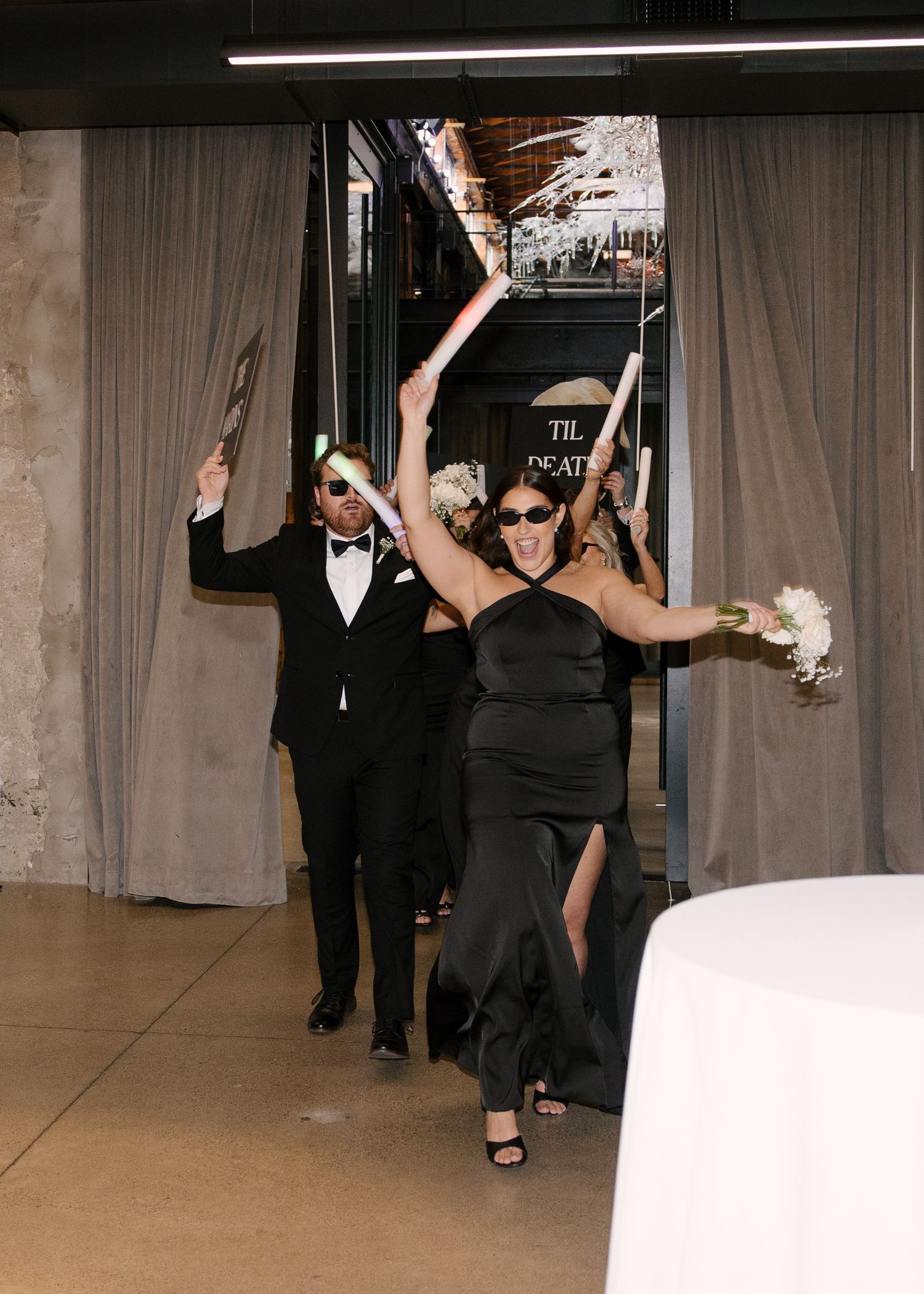Wedding party making grand entrance in black formal attire with raised champagne bottles.