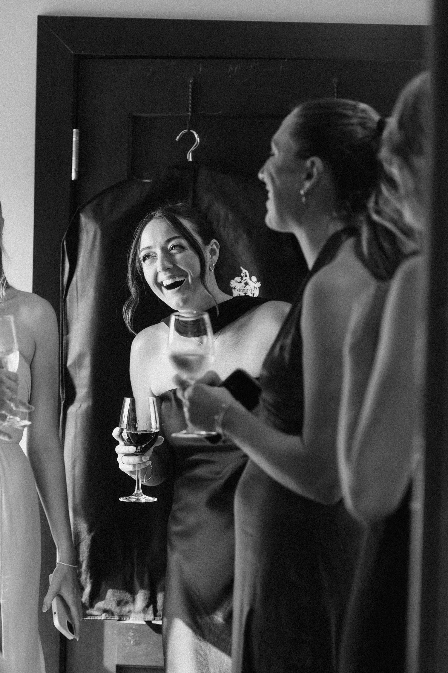 Laughing bridesmaid in satin dress holds wine glass reflected in mirror during wedding preparations.