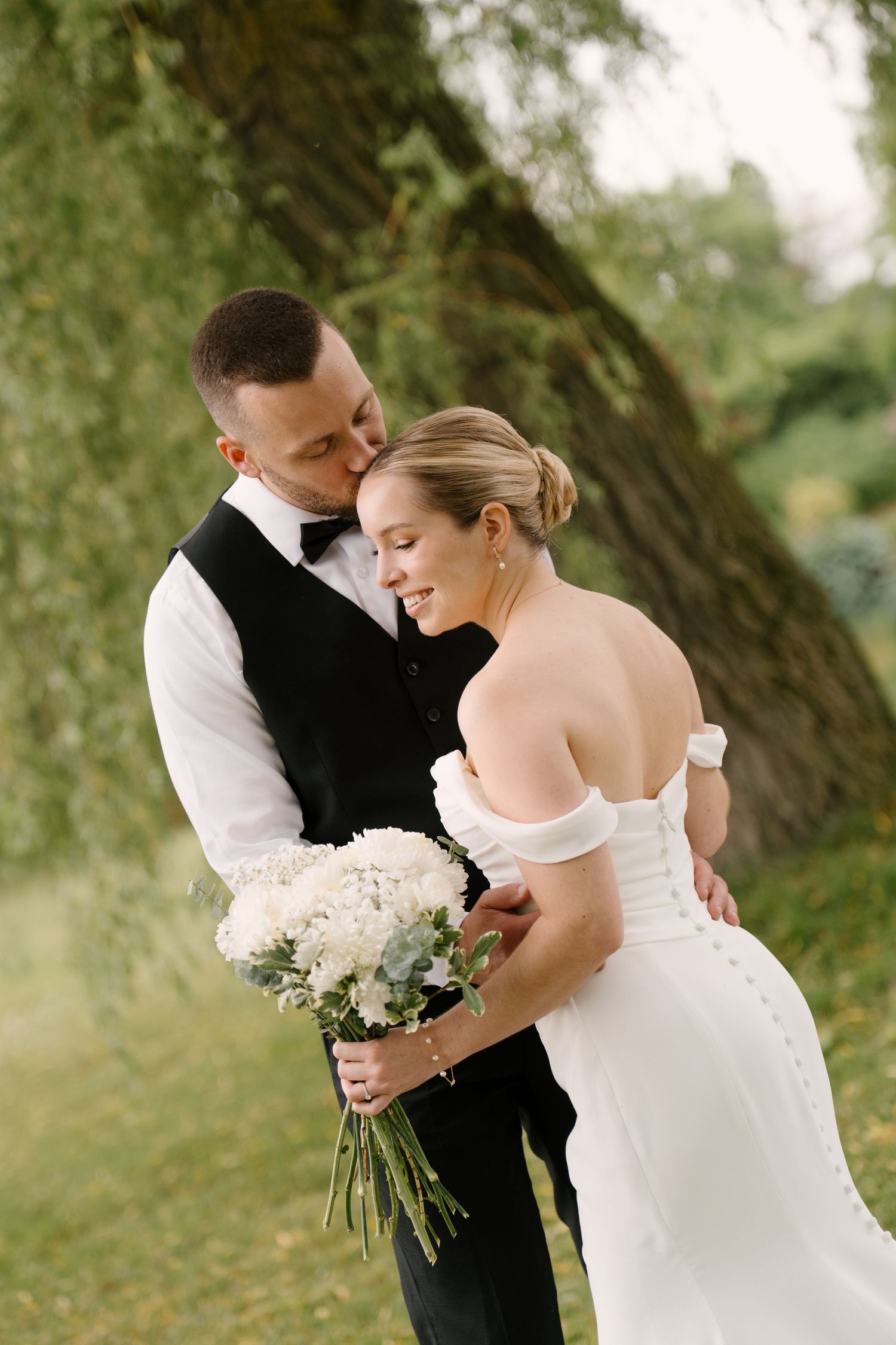 Groom tenderly kisses bride's cheek as she smiles holding white bouquet under willow tree at Guelph golf club.