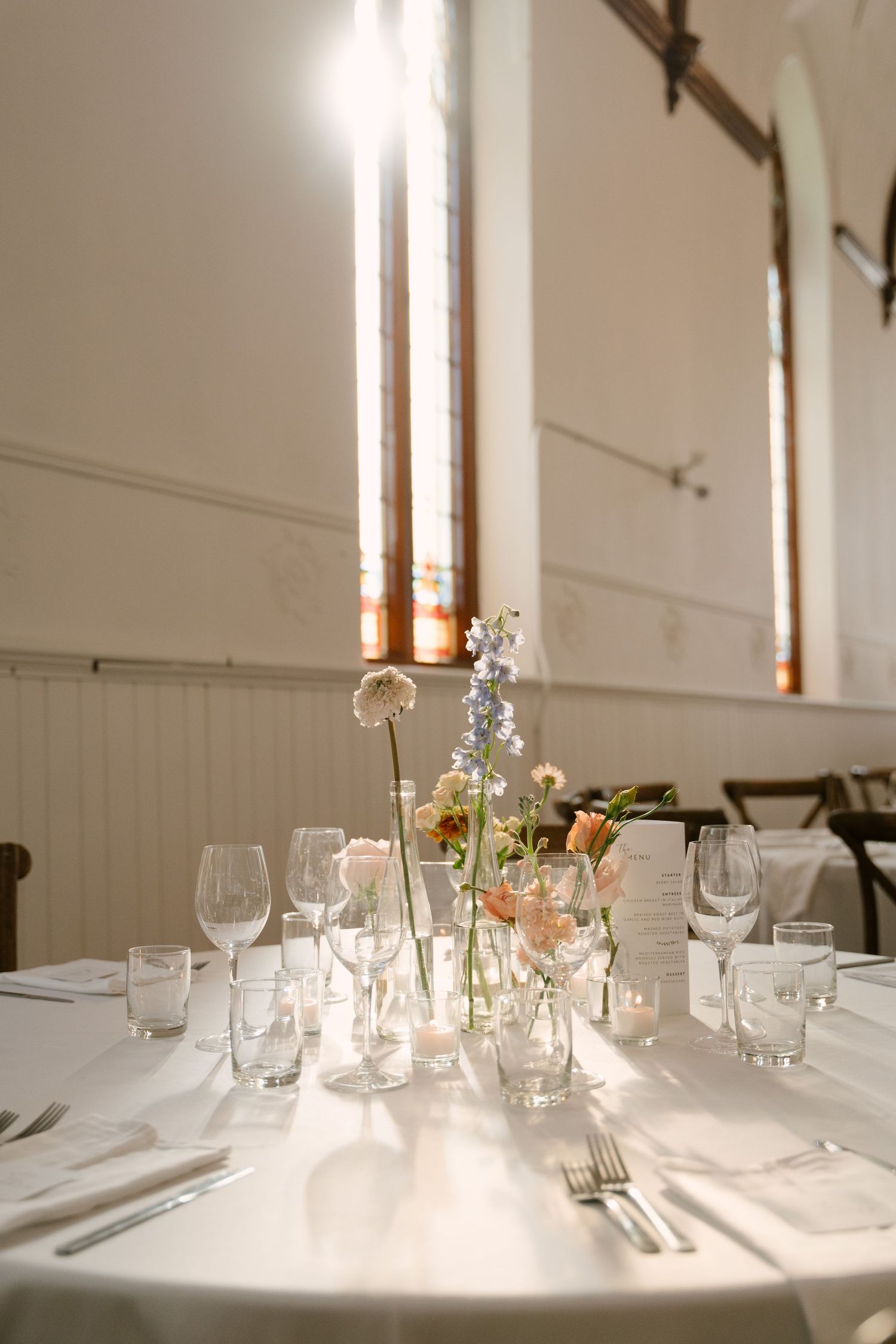 Elegant wedding reception table with floral centerpieces, crystal glassware, and white linens in a sunlit venue.