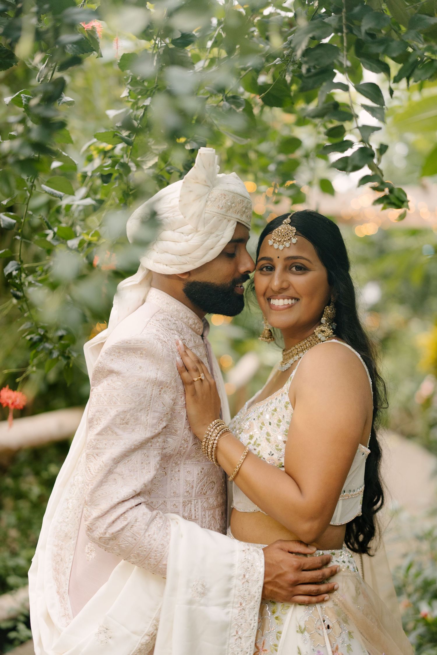 Groom in sherwani kisses smiling bride in embroidered lehenga amid green garden foliage.