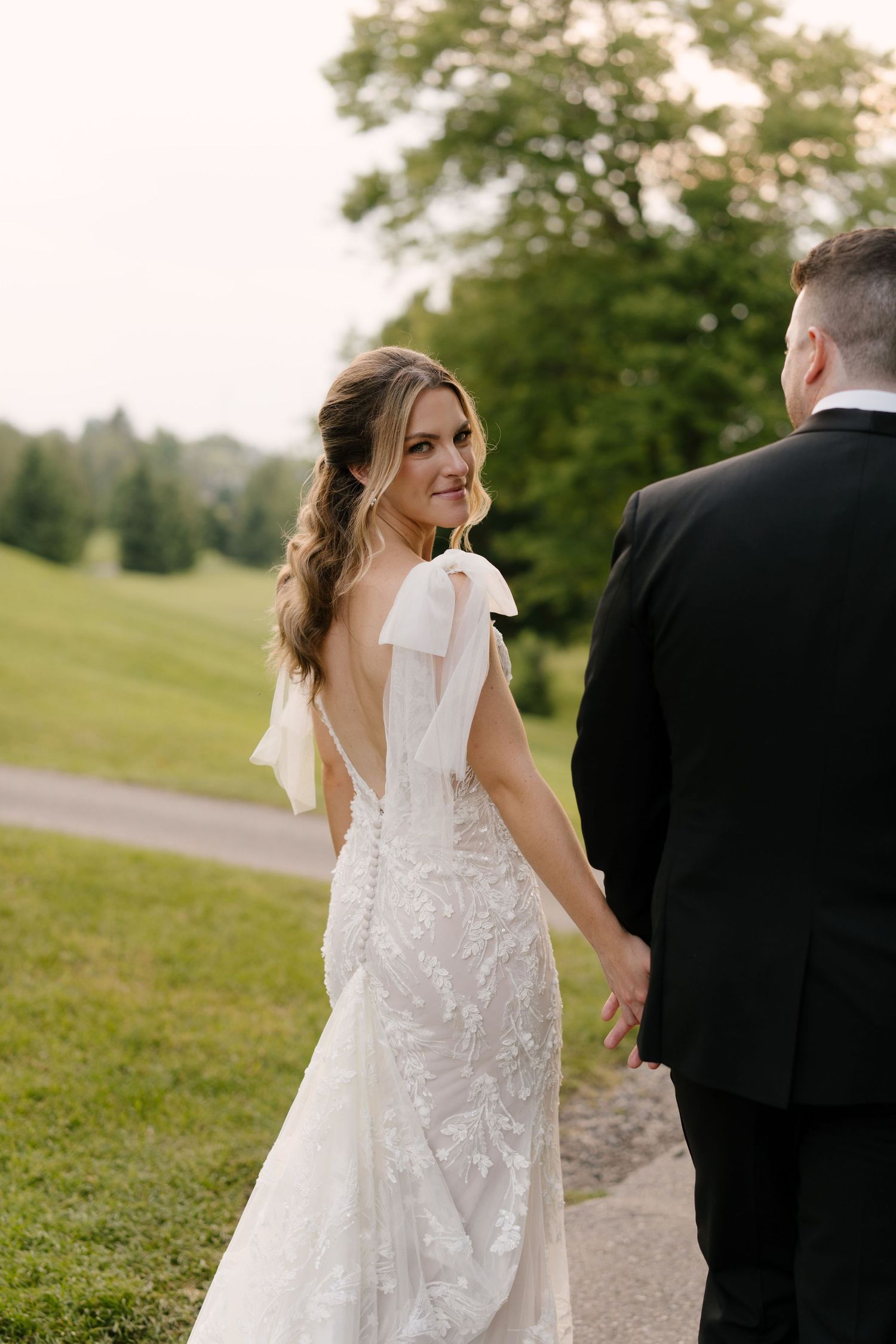 Bride in lace backless gown with bow looks over shoulder while holding groom's hand on a green outdoor path at St Marys Venue