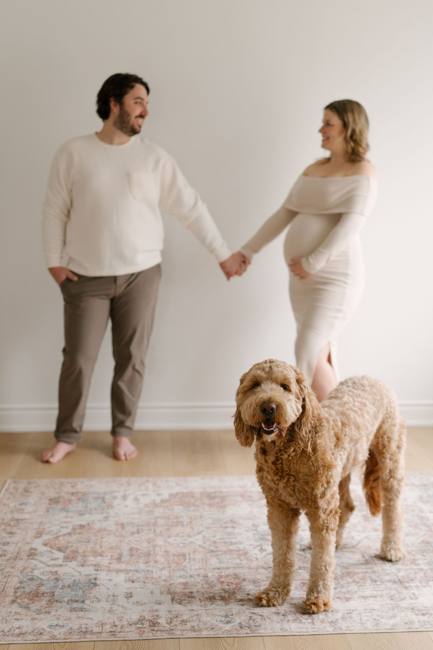Pregnant couple holding hands with a fluffy golden doodle dog standing on a rug in a bright room.