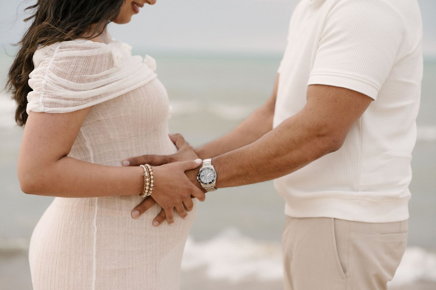Close-up of couple holding hands over pregnant belly on beach, woman wearing pearl bracelet and white dress.