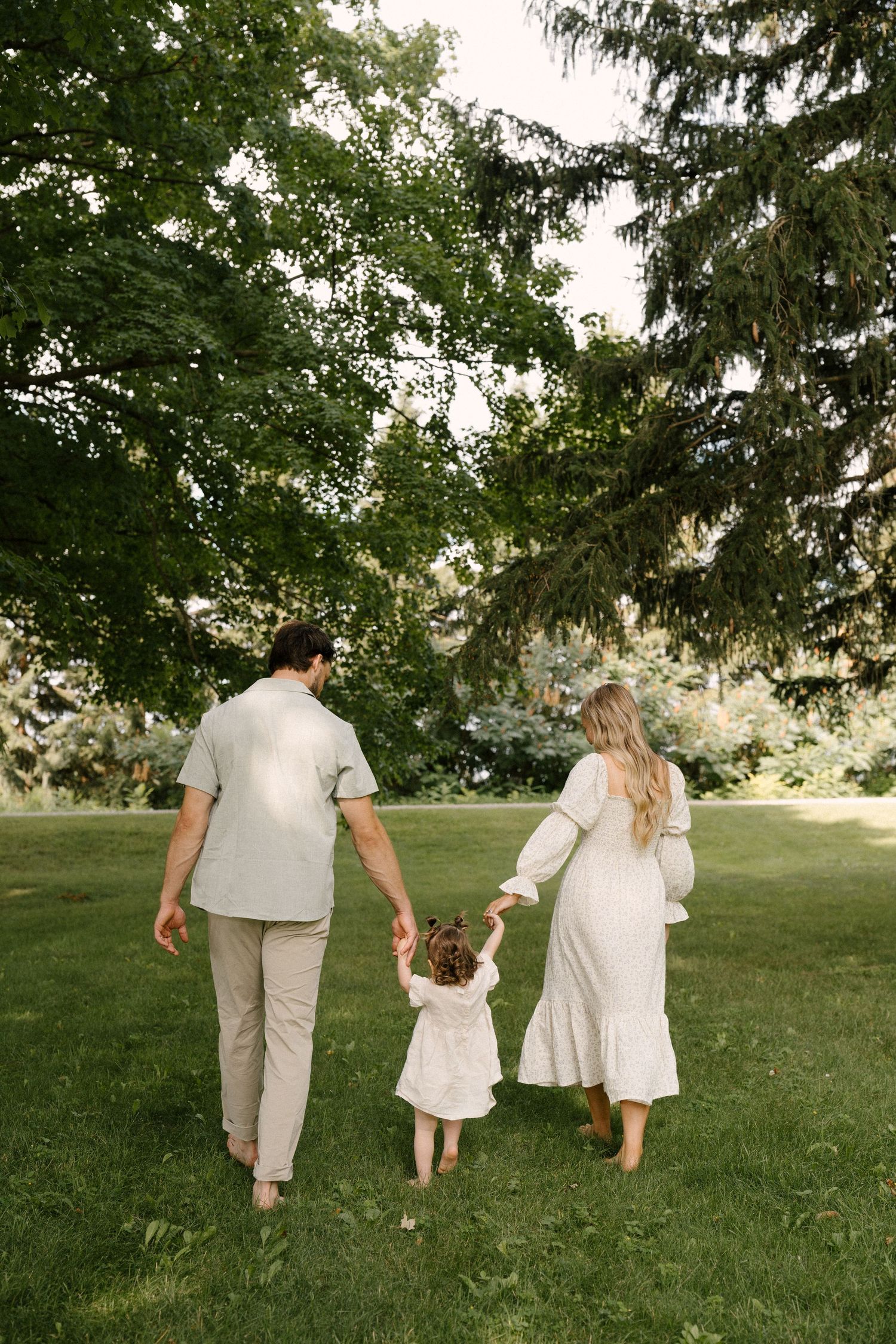 Family of three walking barefoot on green grass holding hands under large trees.