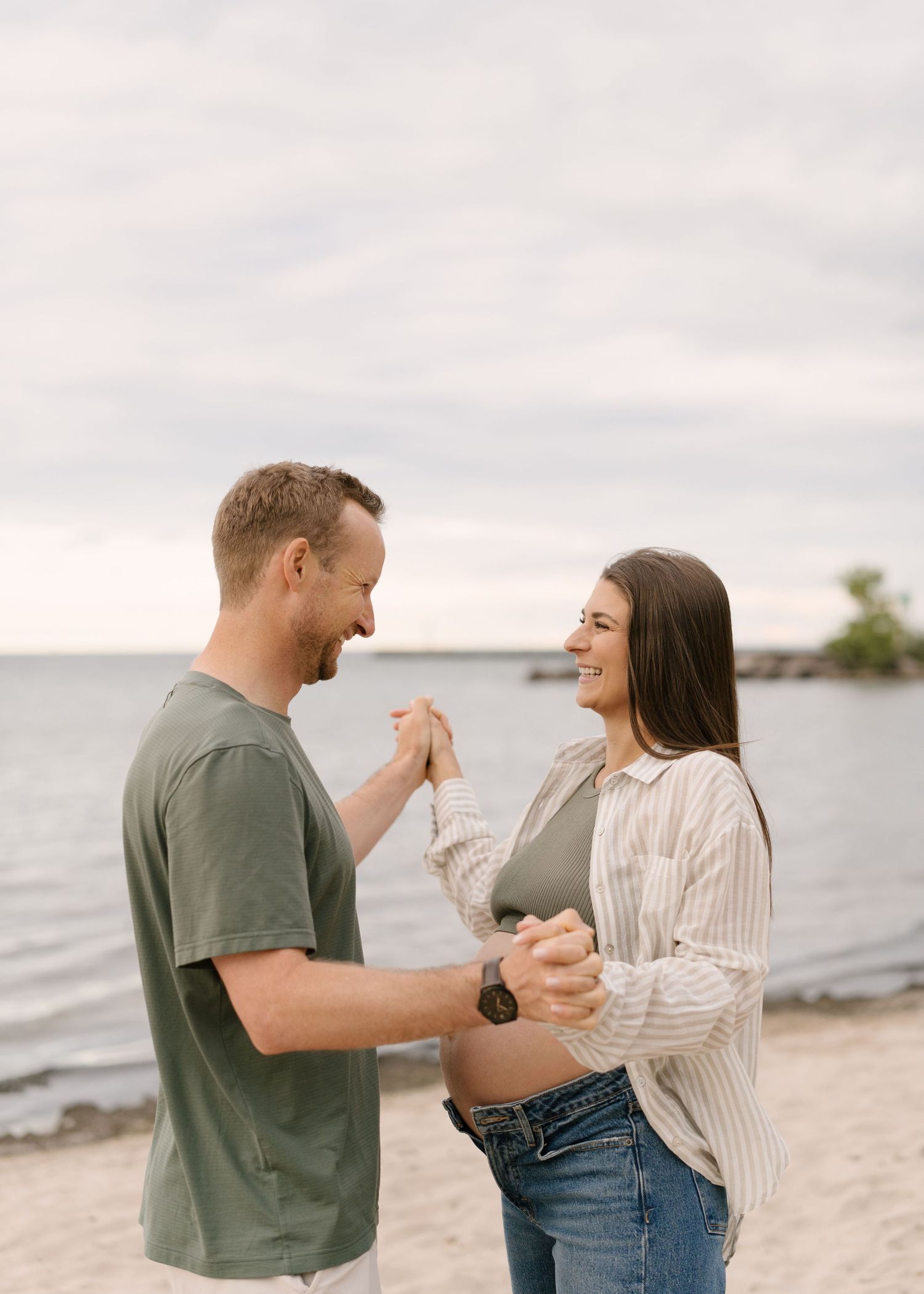 Happy expecting couple holds hands and dances together on a sandy beach with calm water in the background.