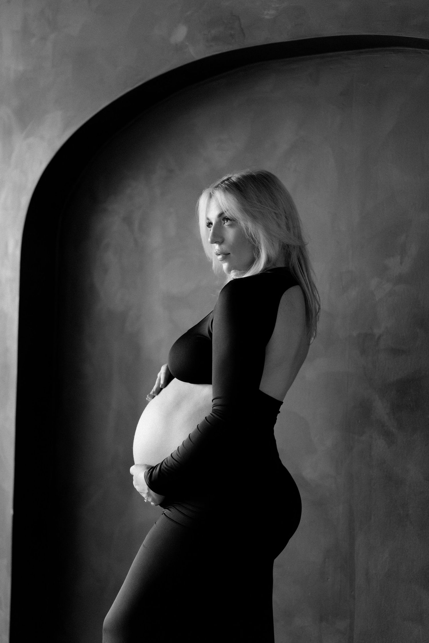 Black and white side profile of pregnant blonde woman in black dress cradling baby bump in arched studio.