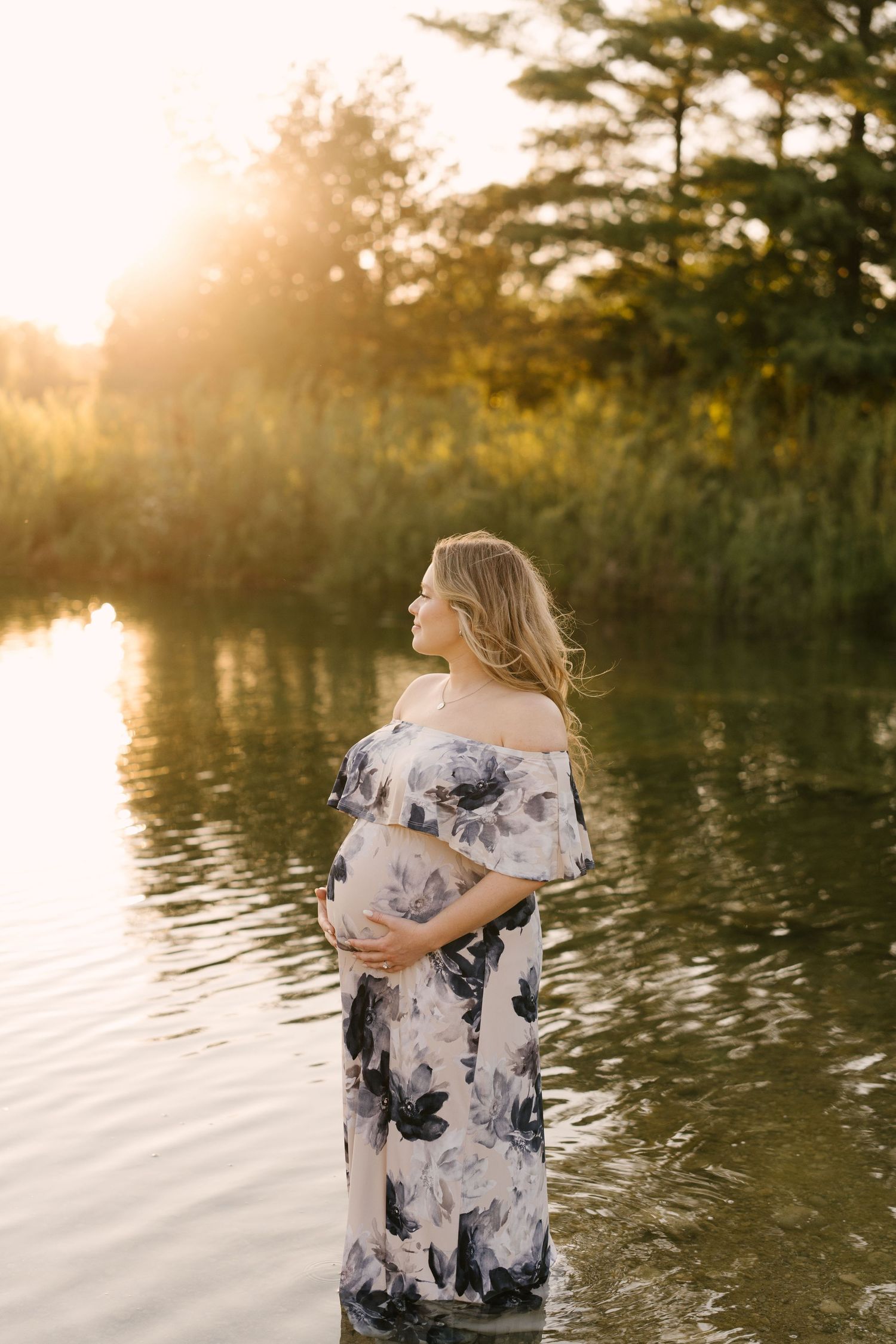 Pregnant woman in floral off-shoulder gown stands in a sunlit lake, cradling her baby bump at sunset.