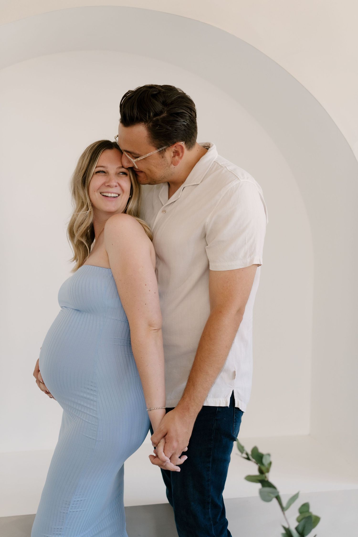 Couple holding hands under arch, man kissing laughing pregnant womans head as she wears a blue dress.