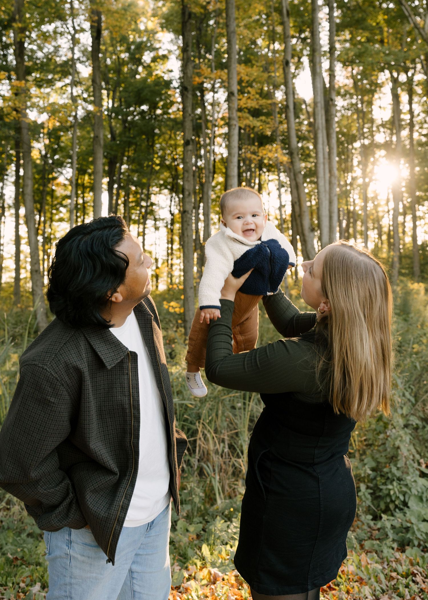 Parents lifting laughing baby in sunlit autumn forest, golden light filtering through trees.
