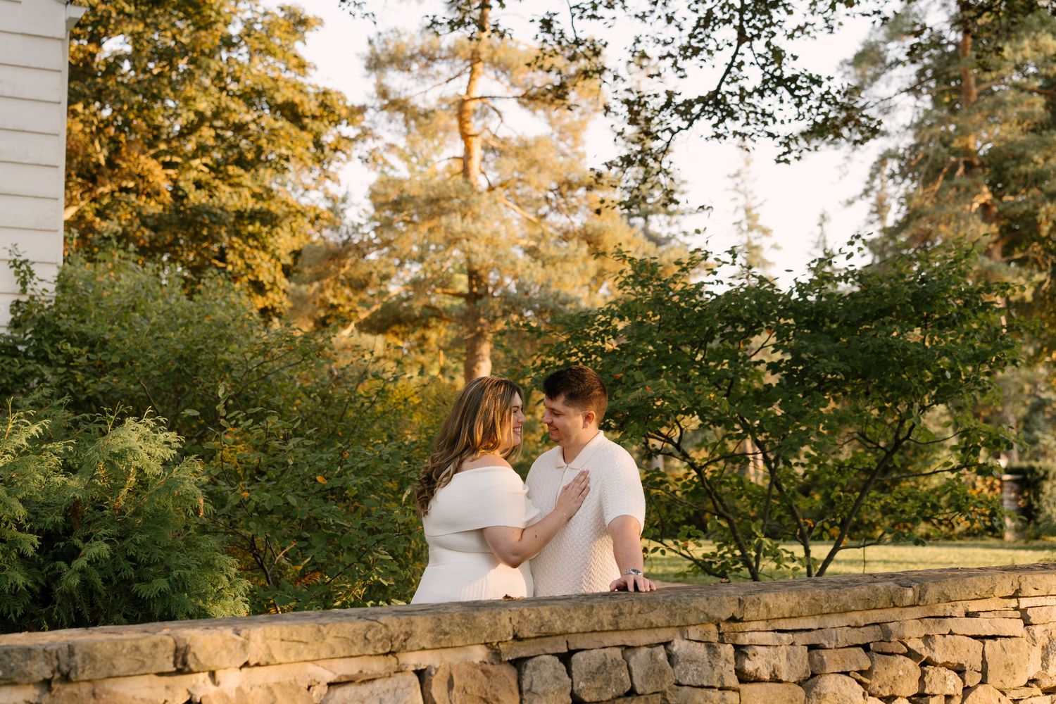 Romantic couple facing each other on stone wall surrounded by lush green trees at sunset.