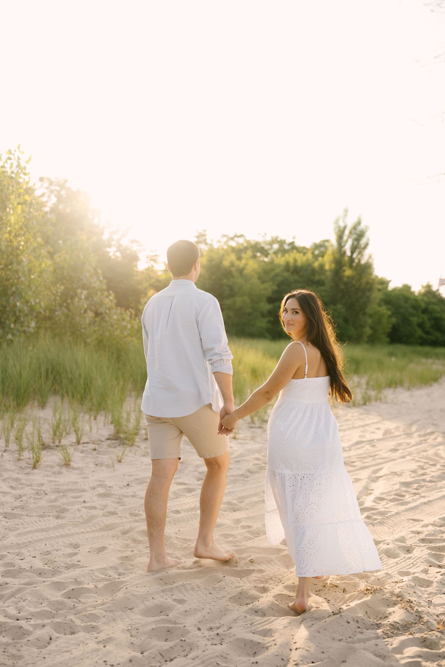 Couple holding hands on sandy beach at sunset, woman in white dress looks back smiling, trees in background.