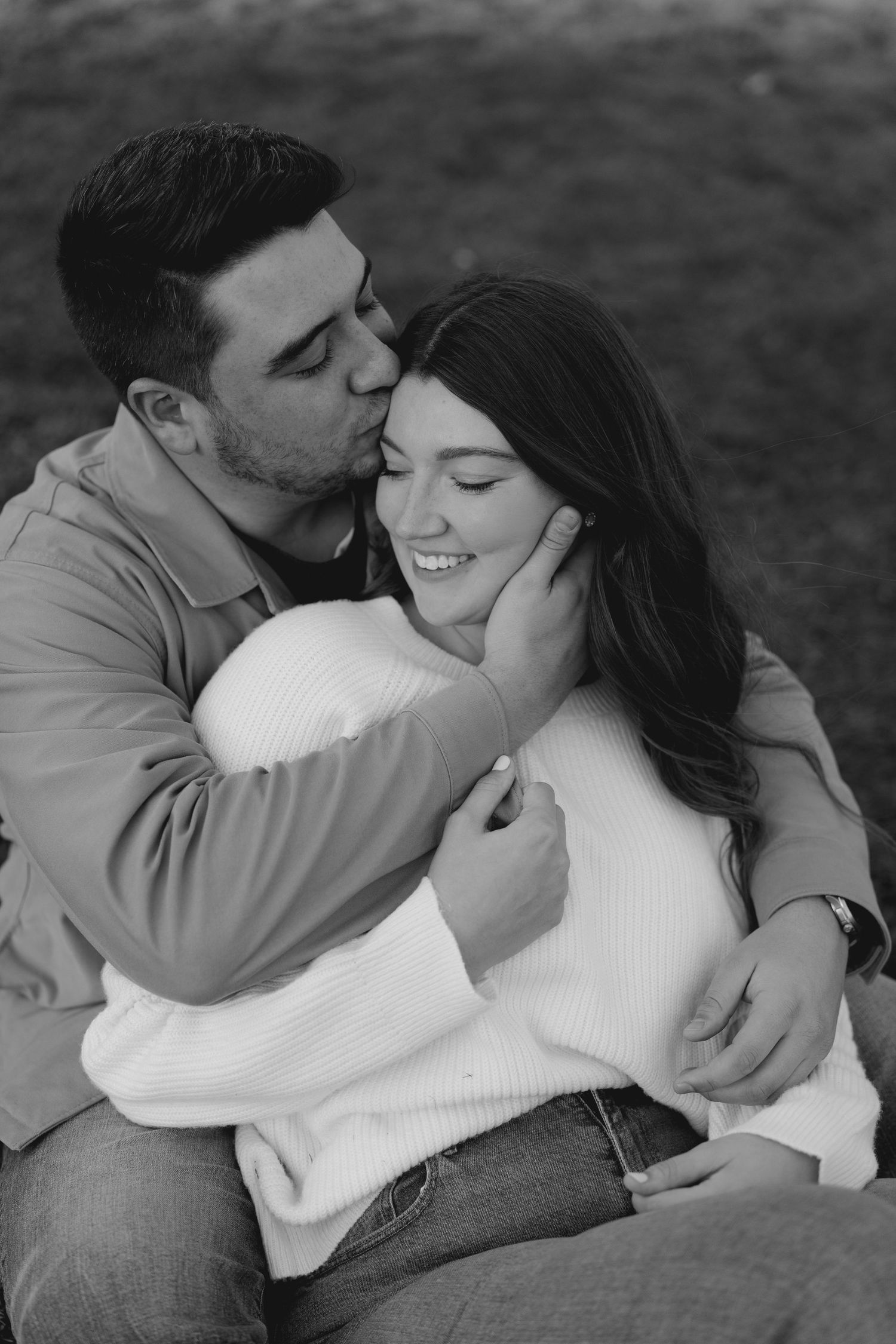 Black and white close-up of couple sitting on grass, man kissing smiling womans cheek tenderly.