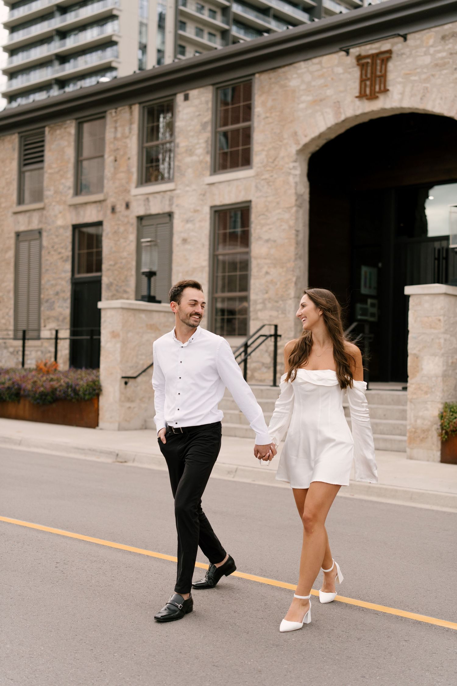 Happy couple holding hands and walking across a street near a brick building.