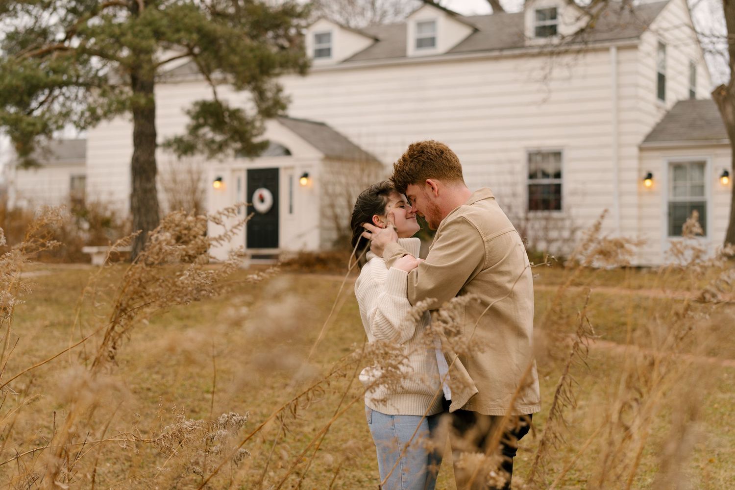 Couple sharing a kiss in a golden meadow with a charming white farmhouse in the background.