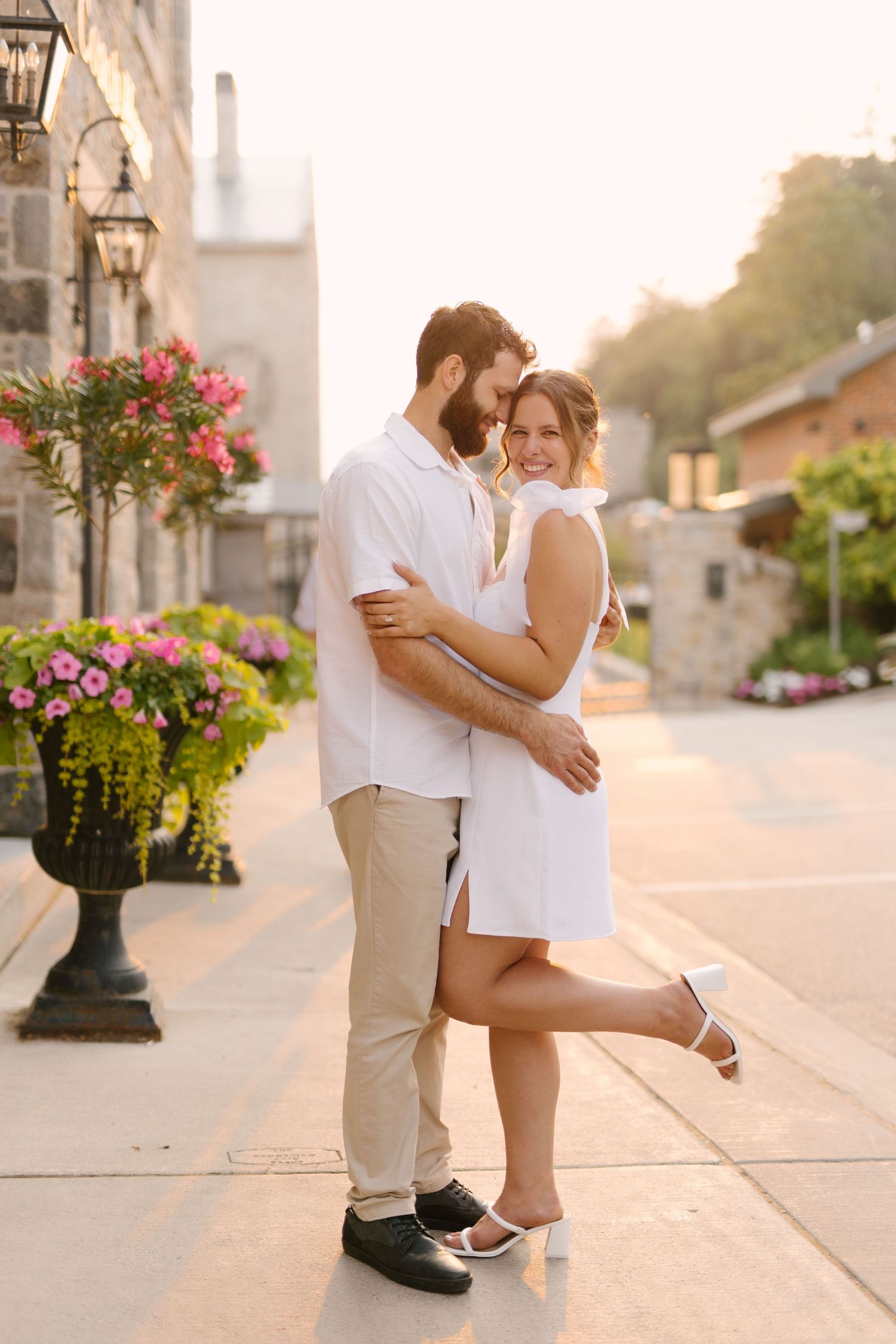 Romantic couple in white outfits embracing on a sunny cobblestone street surrounded by colorful flowers.