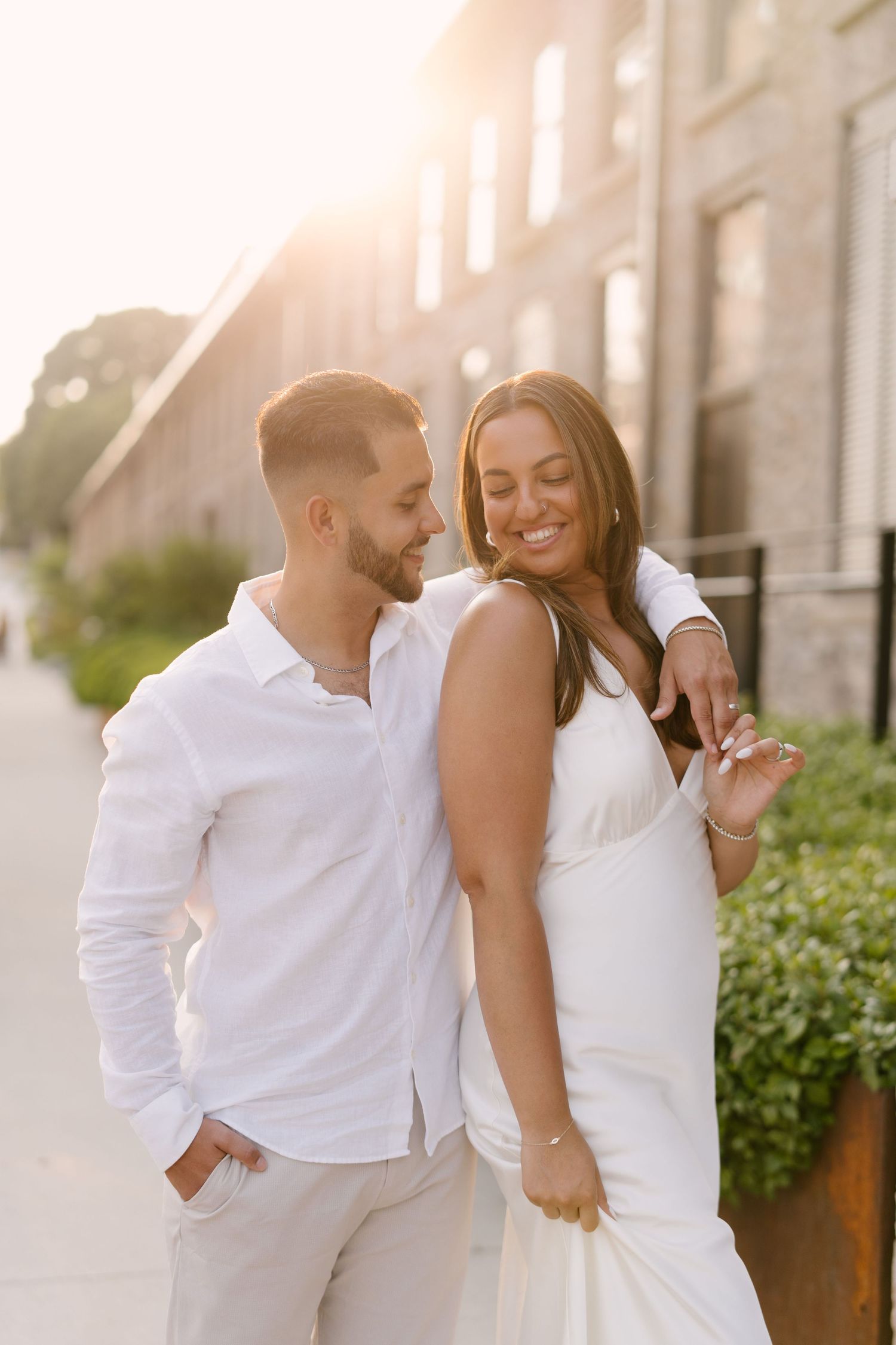 Couple in white outfits embracing during golden hour sunset on urban sidewalk.